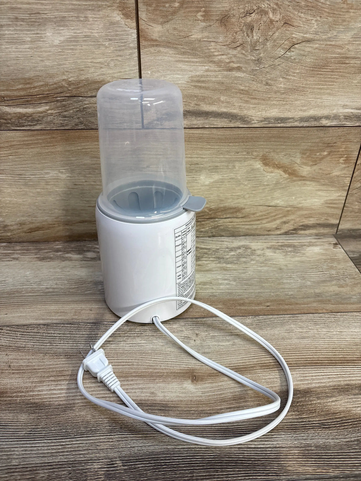Three-quarter view of white Grownsy baby bottle warmer with transparent chamber and power cord on wood surface