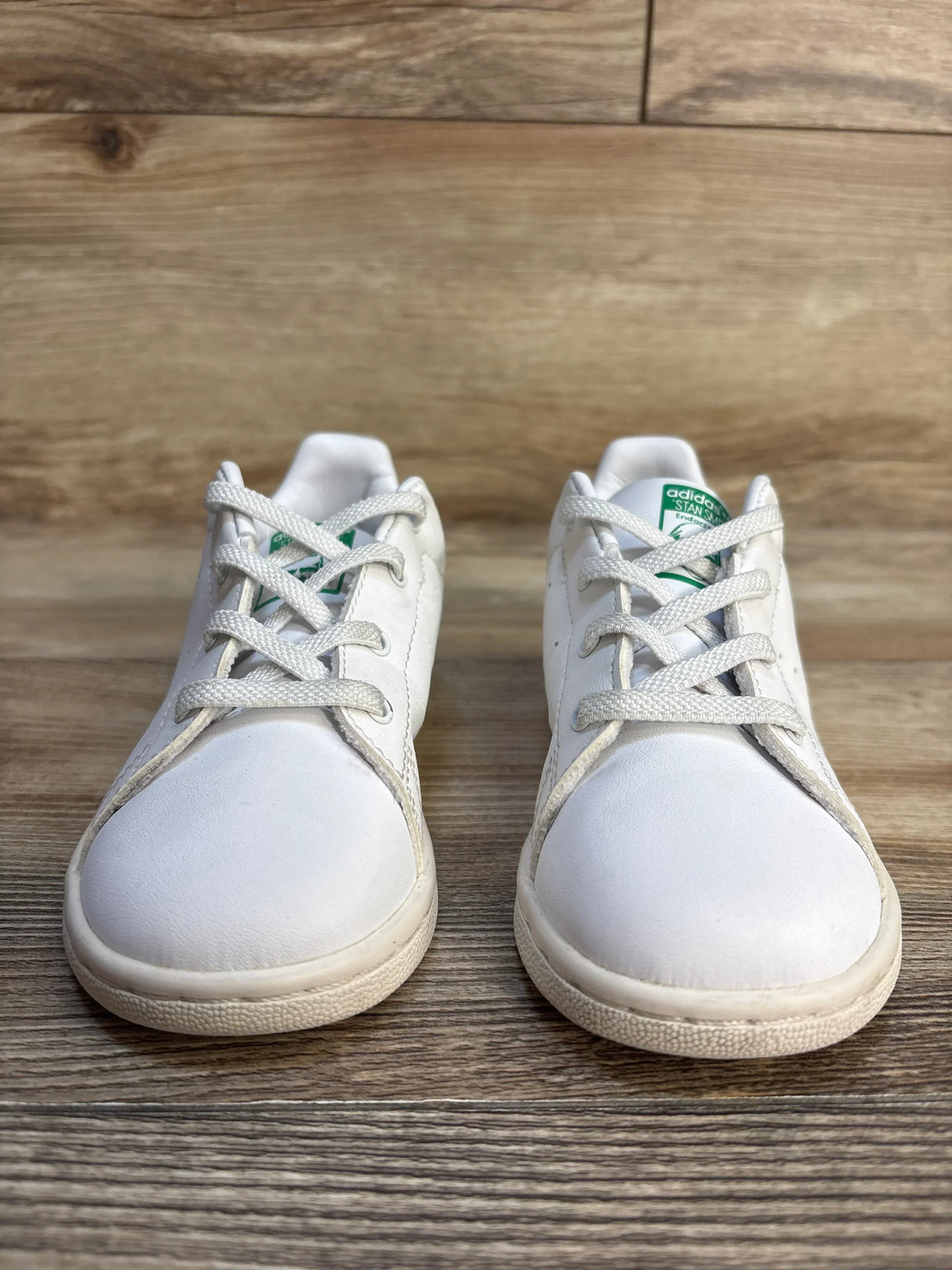 Pair of white Adidas Stan Smith kids sneakers with green logo on wood floor
