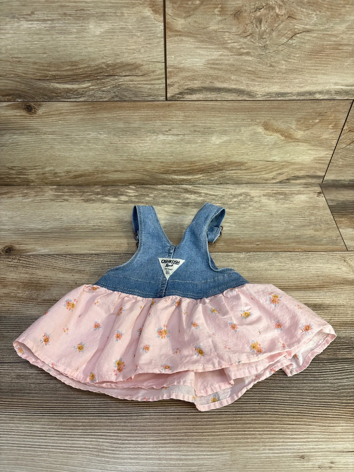 Front flat lay of Oshkosh denim skirtall with pink floral ruffled skirt and triangle logo patch on wooden floor