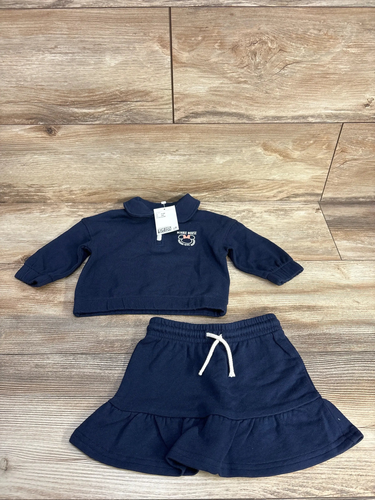 Flat lay of NWT navy baby two piece outfit set showcasing long sleeve collared top with embroidered patch and ruffle skirt with white drawstring on wood plank floor