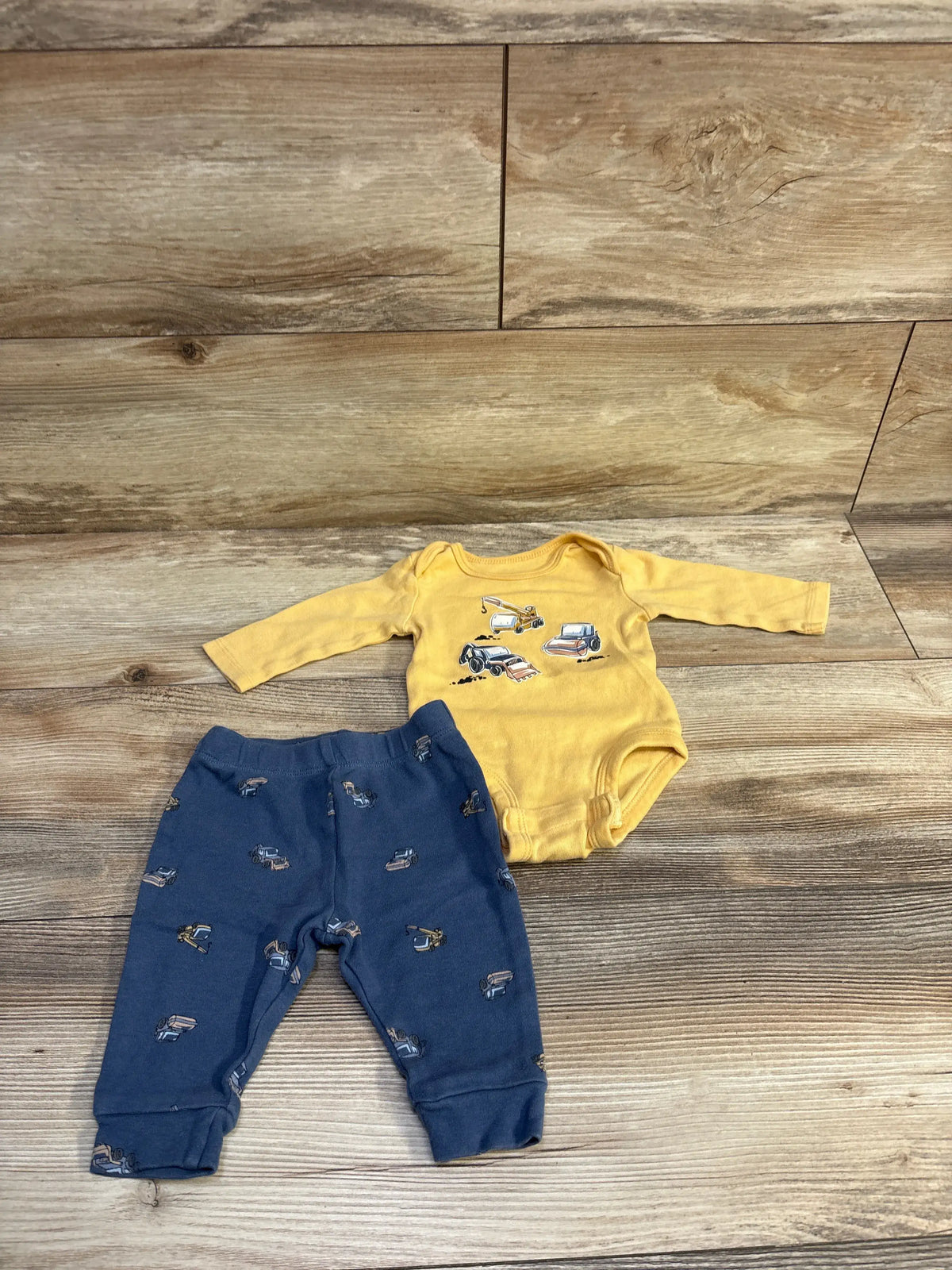 Flat lay of 2-piece baby set: yellow long-sleeve bodysuit with construction vehicle graphics and navy truck-print pants on wood background