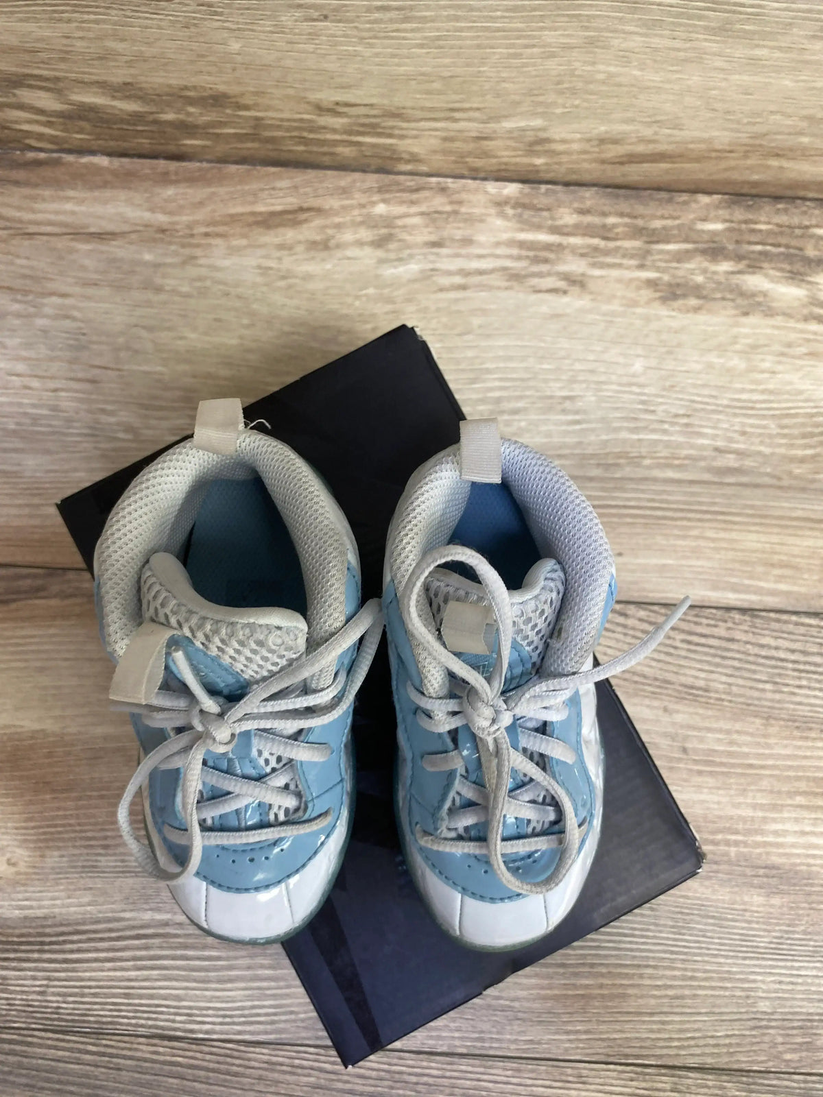 Top-down view of Nike Little Posite One TD toddler sneakers in aura worn blue and white, lace-up with mesh collar on a black shoebox and wood floor