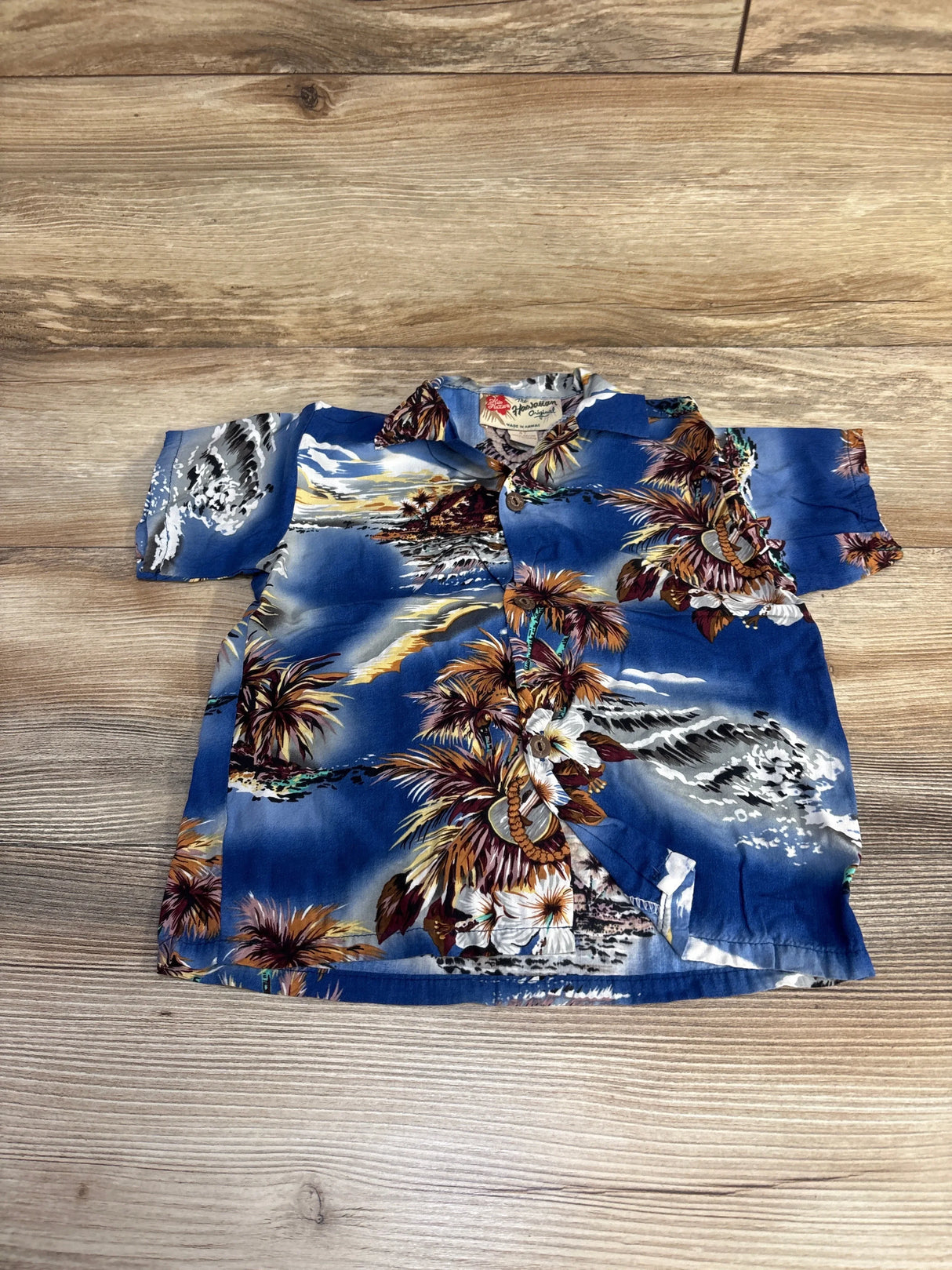 Front view of toddler blue Hilo Hattie aloha button-up shirt showing tropical palm, flower, and island print on wood floor