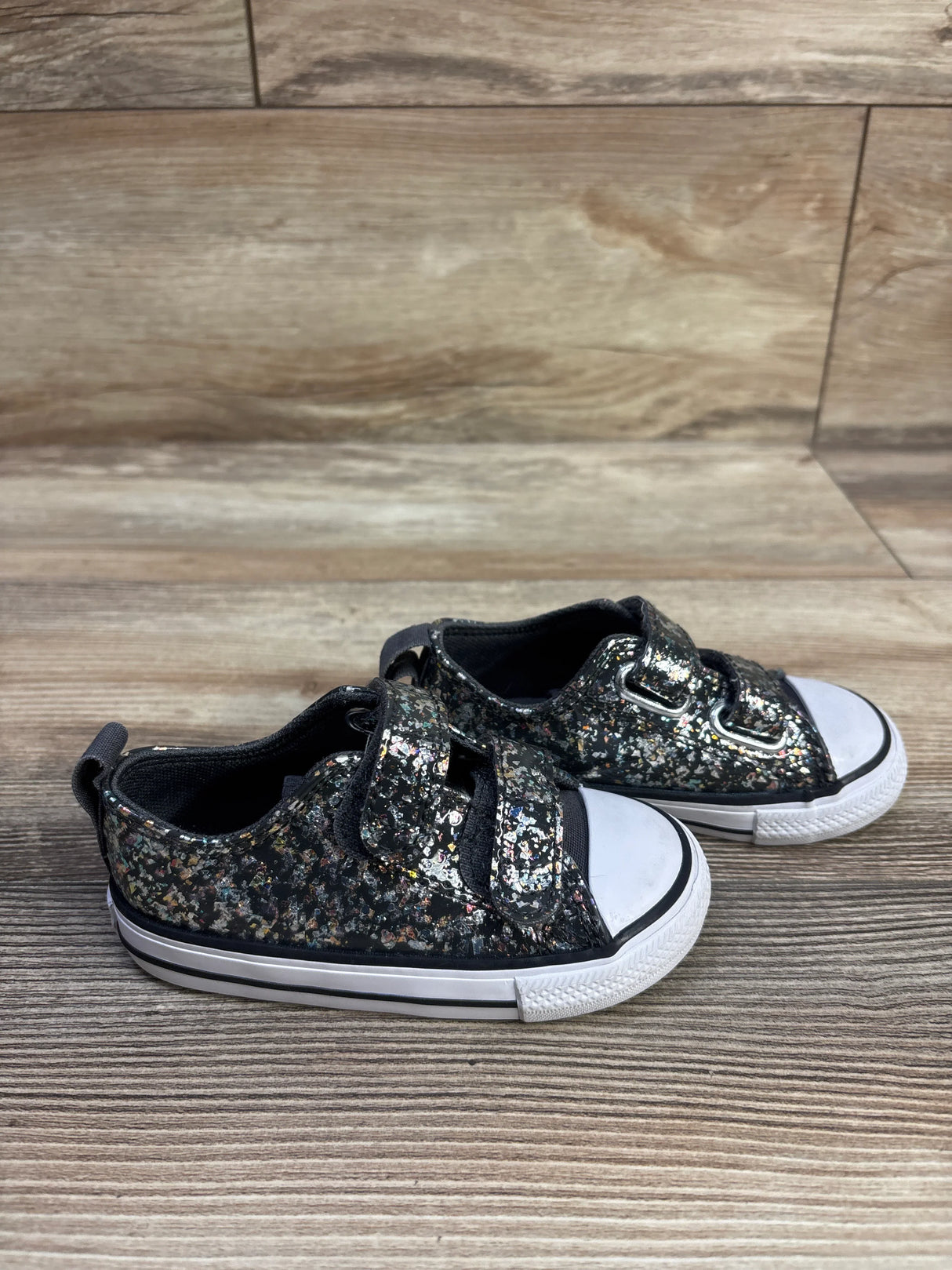 Angled side view of size 6C black glitter Converse Chuck Taylor toddler sneakers with double Velcro straps on a wood floor