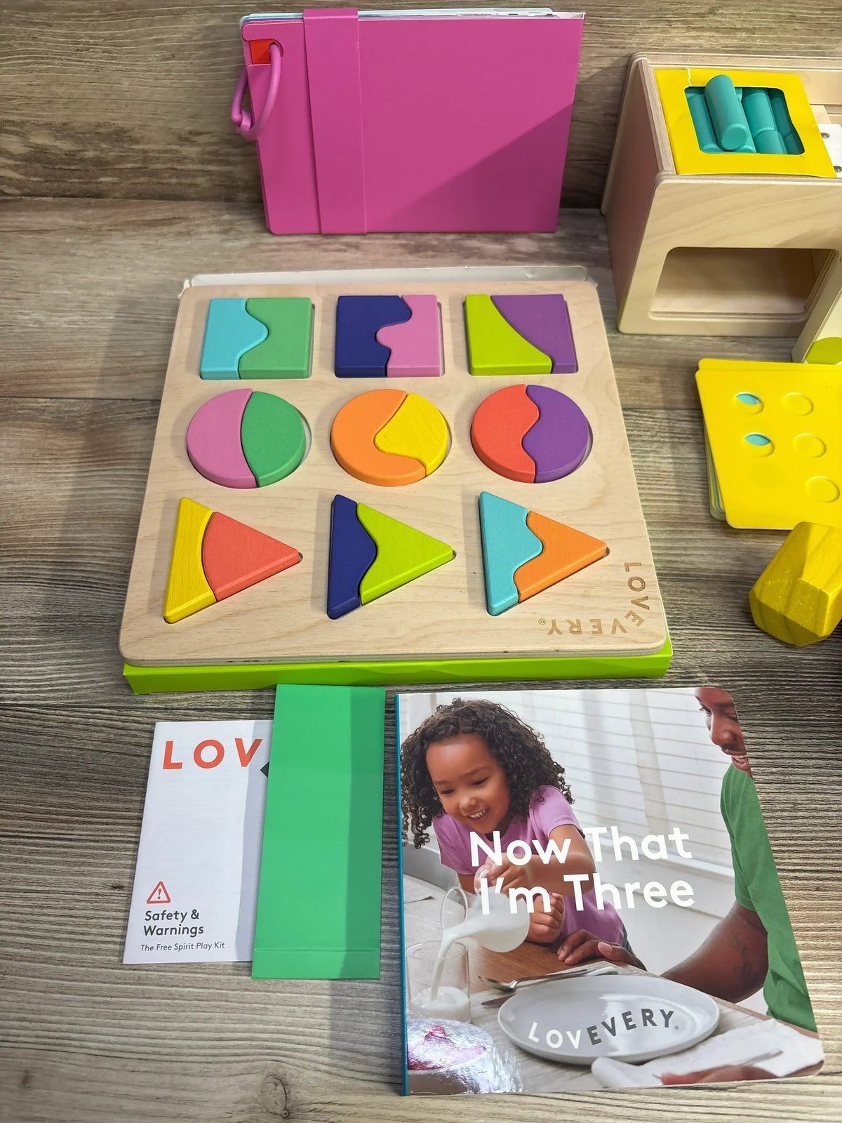 Top-down view of Lovevery Free Spirit Play Kit, wooden multicolor shape-matching board, peg block, and the book ‘Now That I’m Three’ on a wood surface