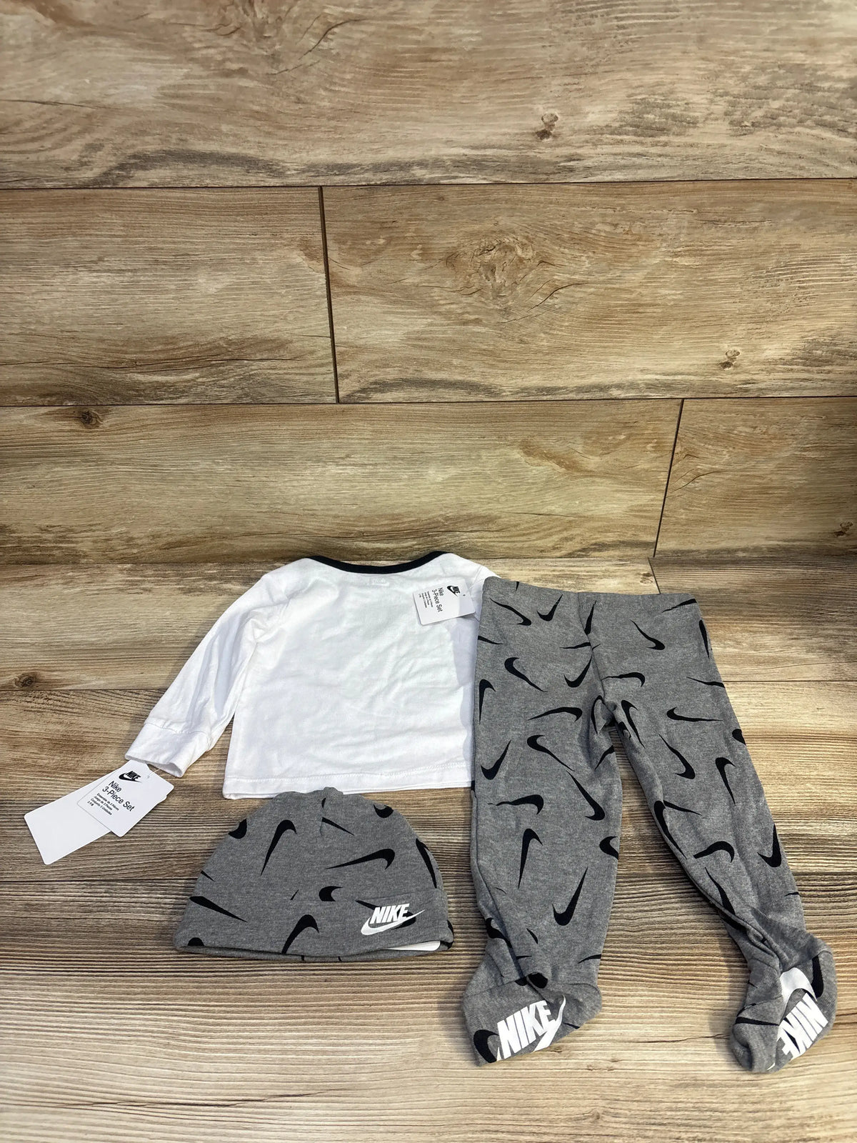 Flat lay of Nike 3pc baby set: white long-sleeve top, grey footed pants and hat with black swoosh print on wood background