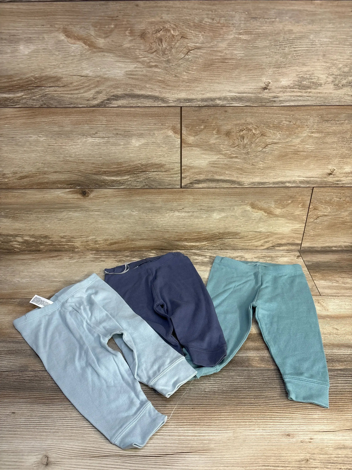 Flat lay of three Cloud Island baby pull-on pants with elastic waist and cuffed ankles in mint, navy, and teal on wood background