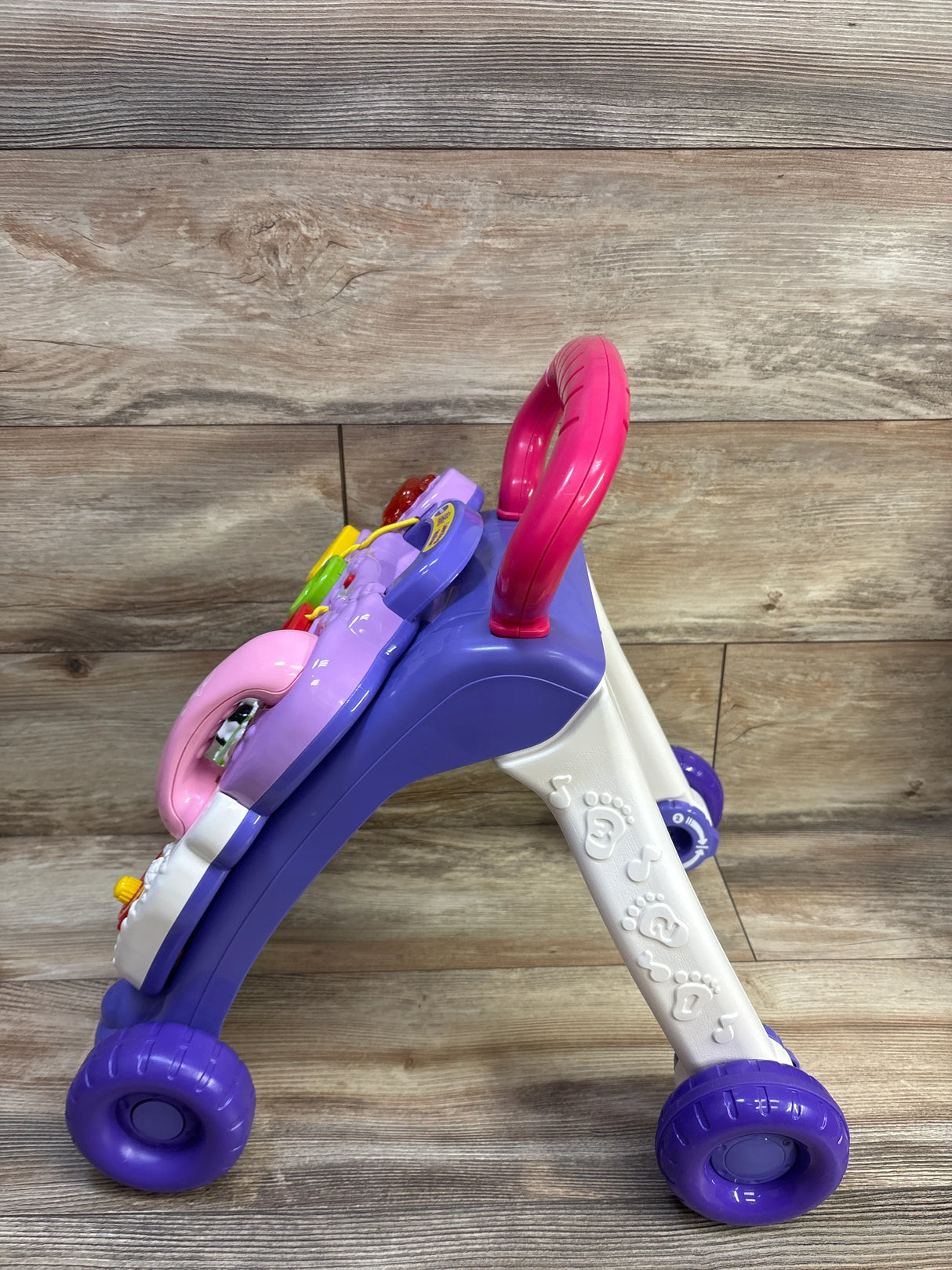 Side view of VTech Sit-to-Stand Learning Walker showing purple frame, pink handle, activity panel and purple wheels on floor