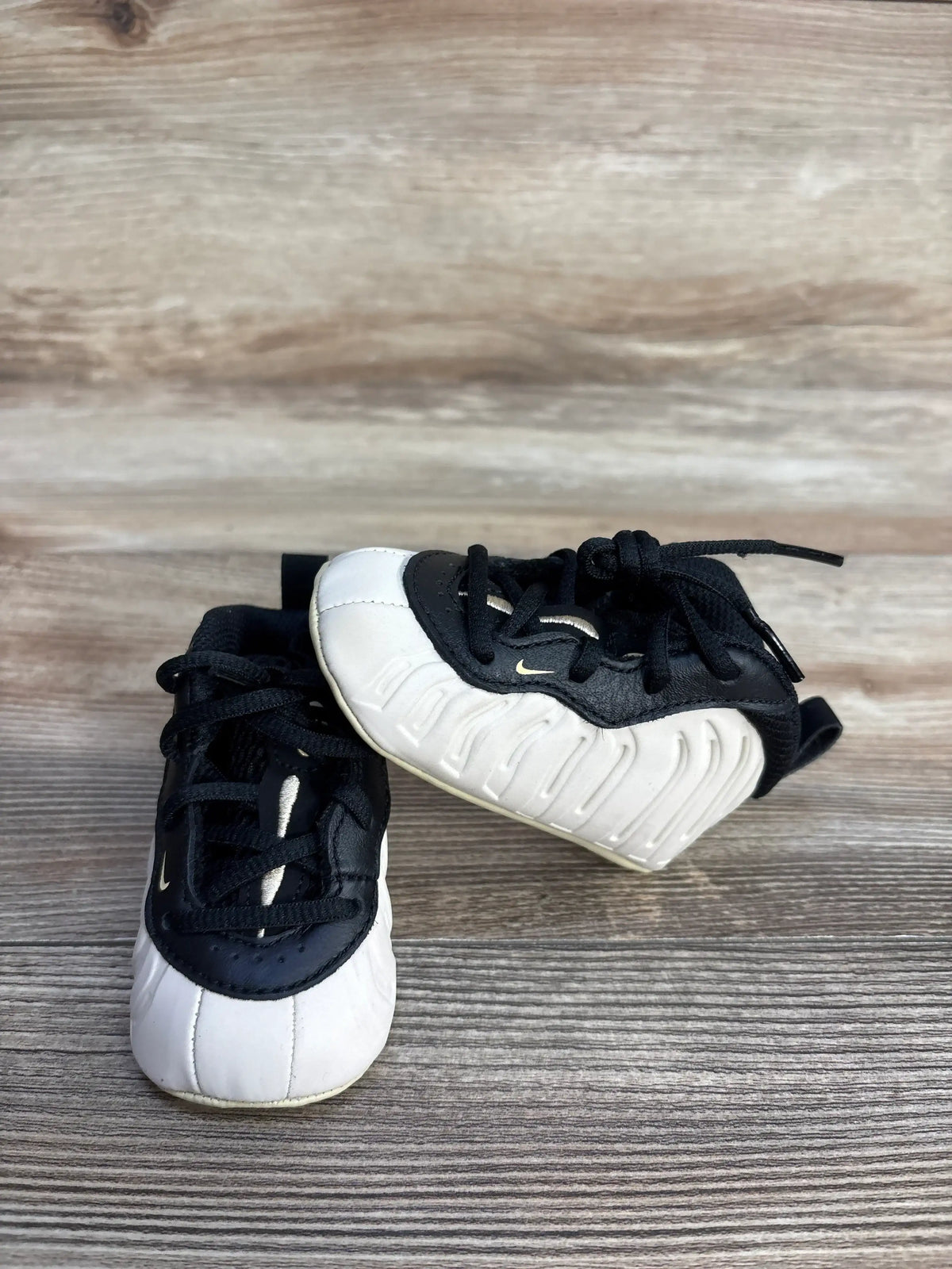 Three-quarter view of Nike Posite One Crib Booties Light Orewood Brown sneakers size 3c in white and black with foamposite upper on wood plank floor
