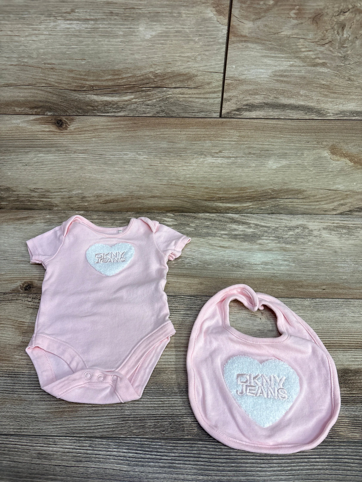 Pink DKNY JEANS baby onesie and matching bib with white heart logo on wooden floor