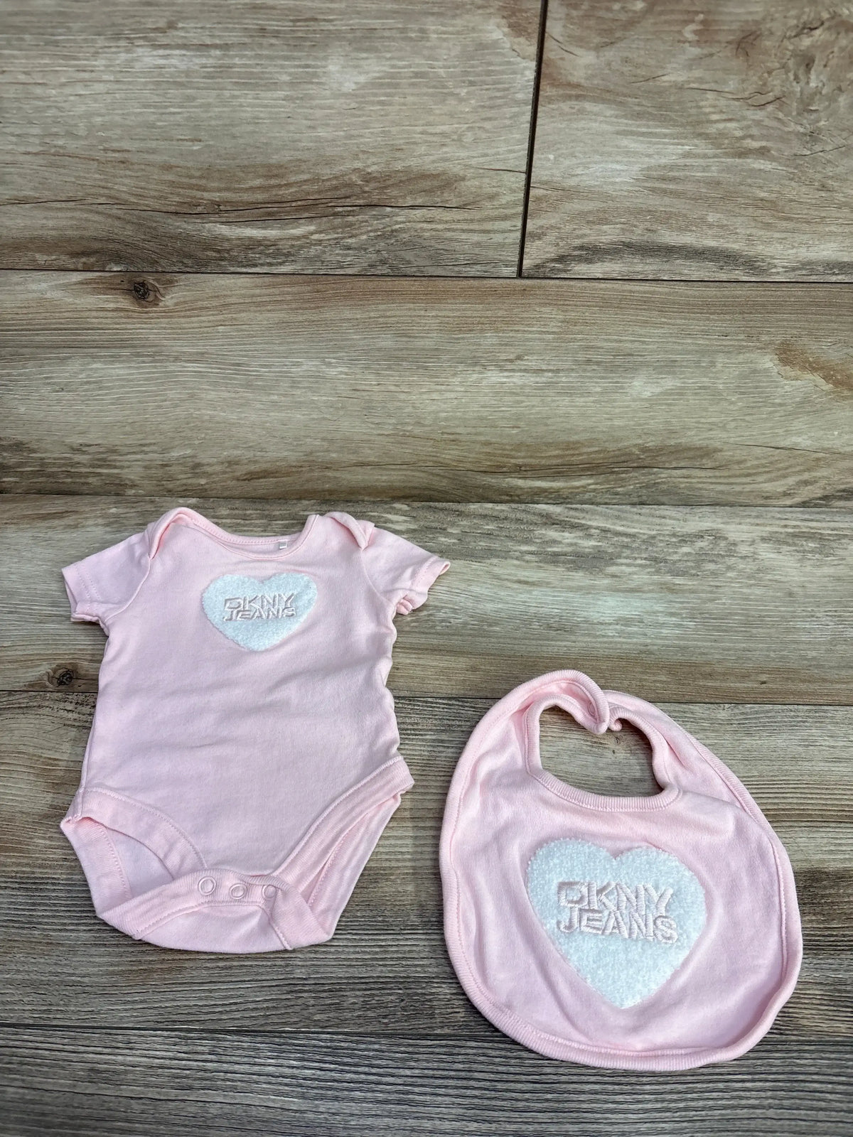 Pink DKNY JEANS baby onesie and matching bib with white heart logo on wooden floor