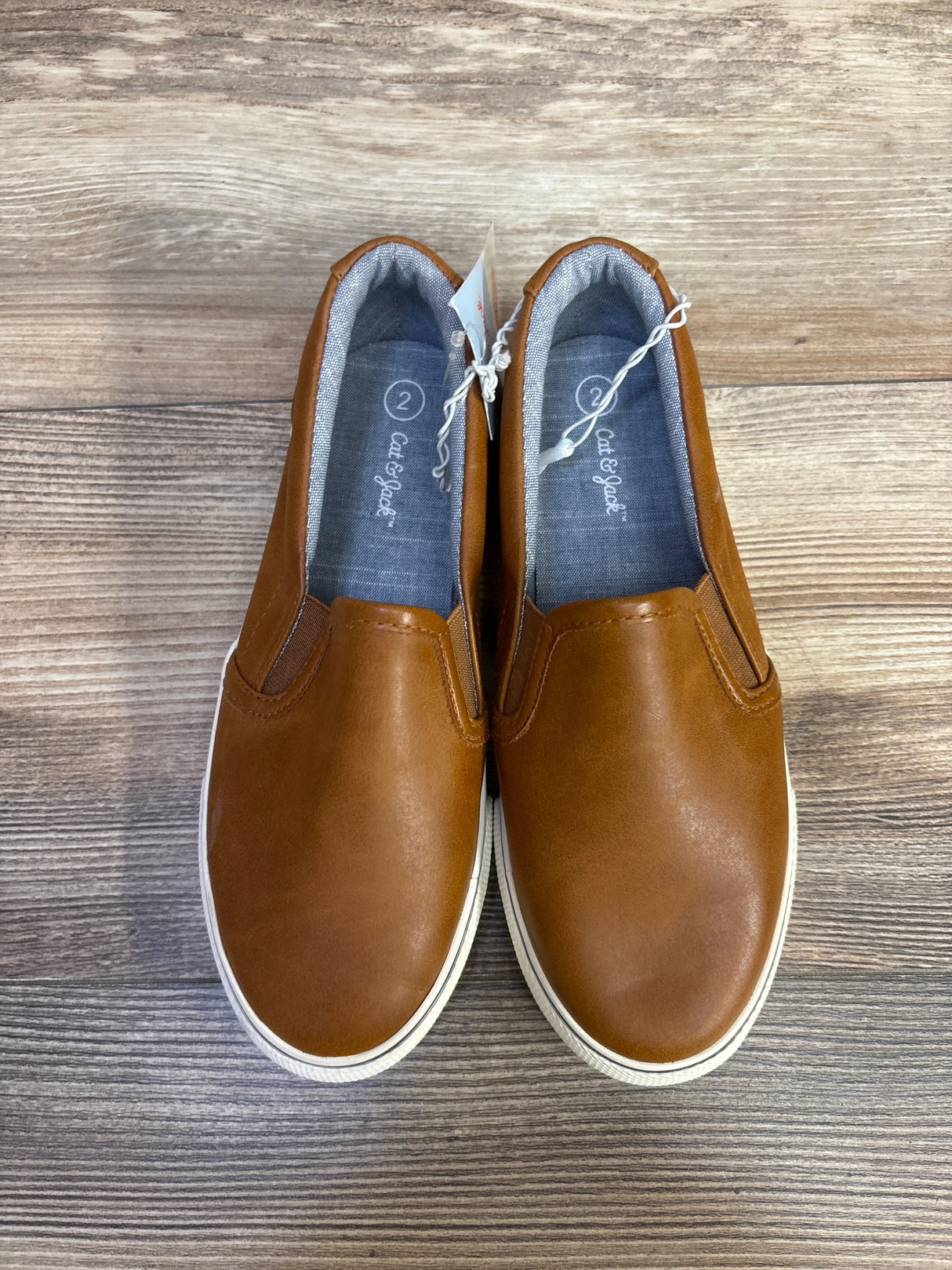 Top-down view of toddler Cat & Jack slip-on sneakers showing brown faux-leather upper, elastic side panels and white rubber sole on wood floor