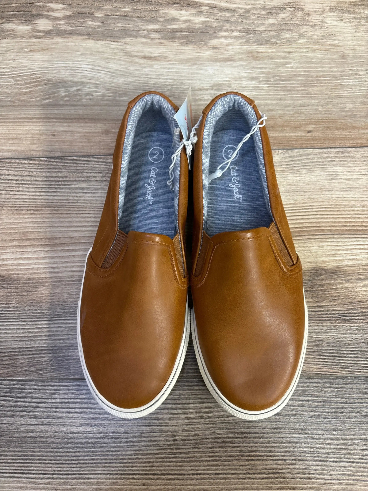 Top-down view of toddler Cat & Jack slip-on sneakers showing brown faux-leather upper, elastic side panels and white rubber sole on wood floor