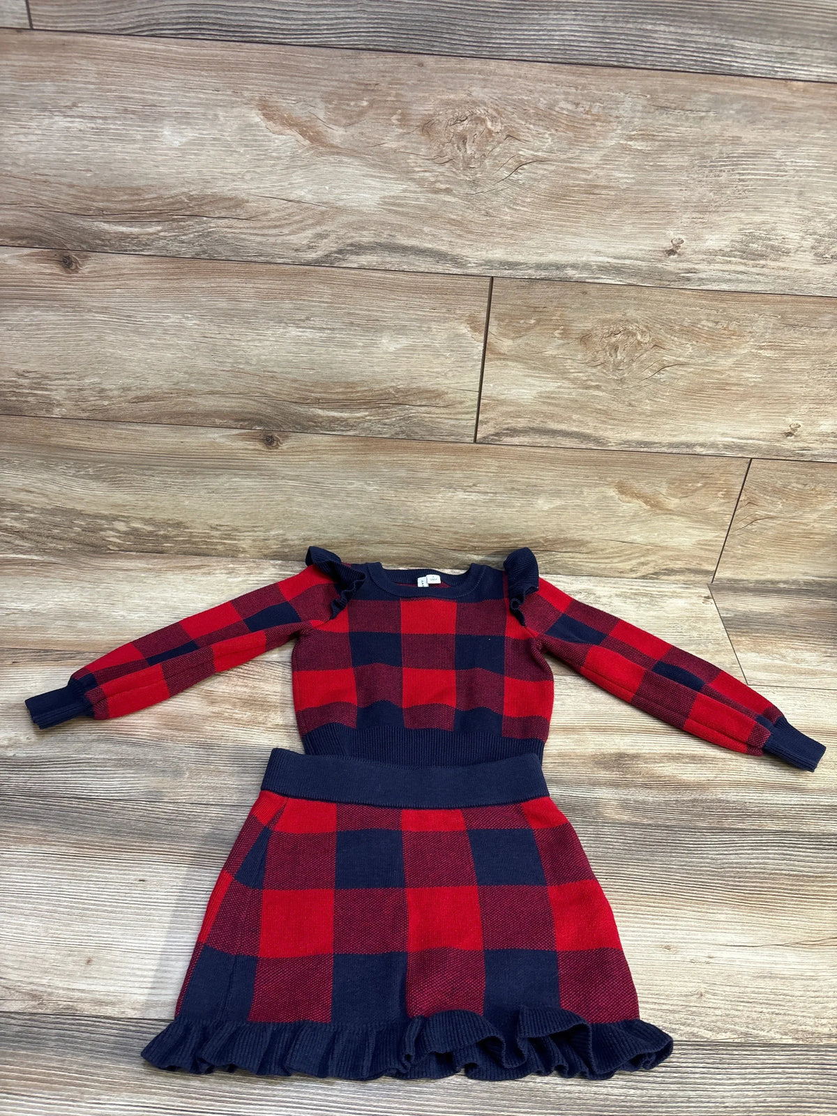 Flat lay of Janie & Jack two-piece plaid set in Fireside red showing ruffle shoulders and ruffled hem on wood floor