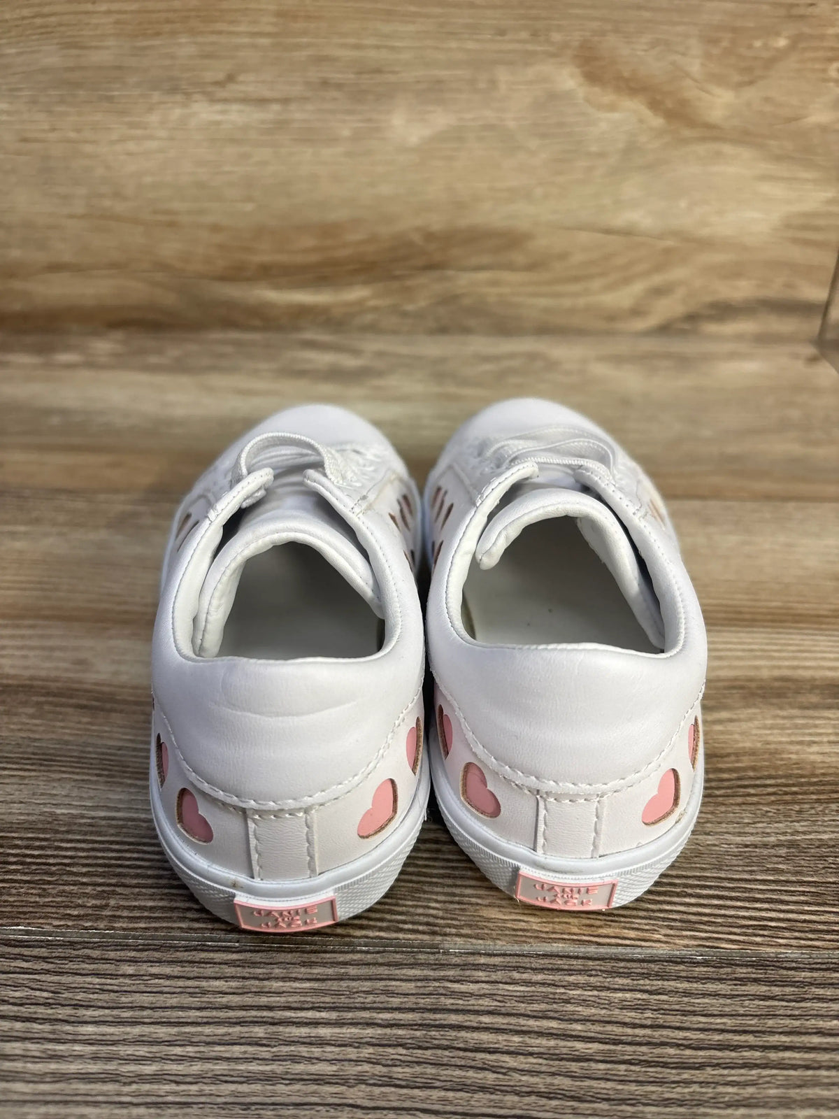 Rear view of white Janie & Jack heart sneakers showcasing pink heart details on a wood-grain background