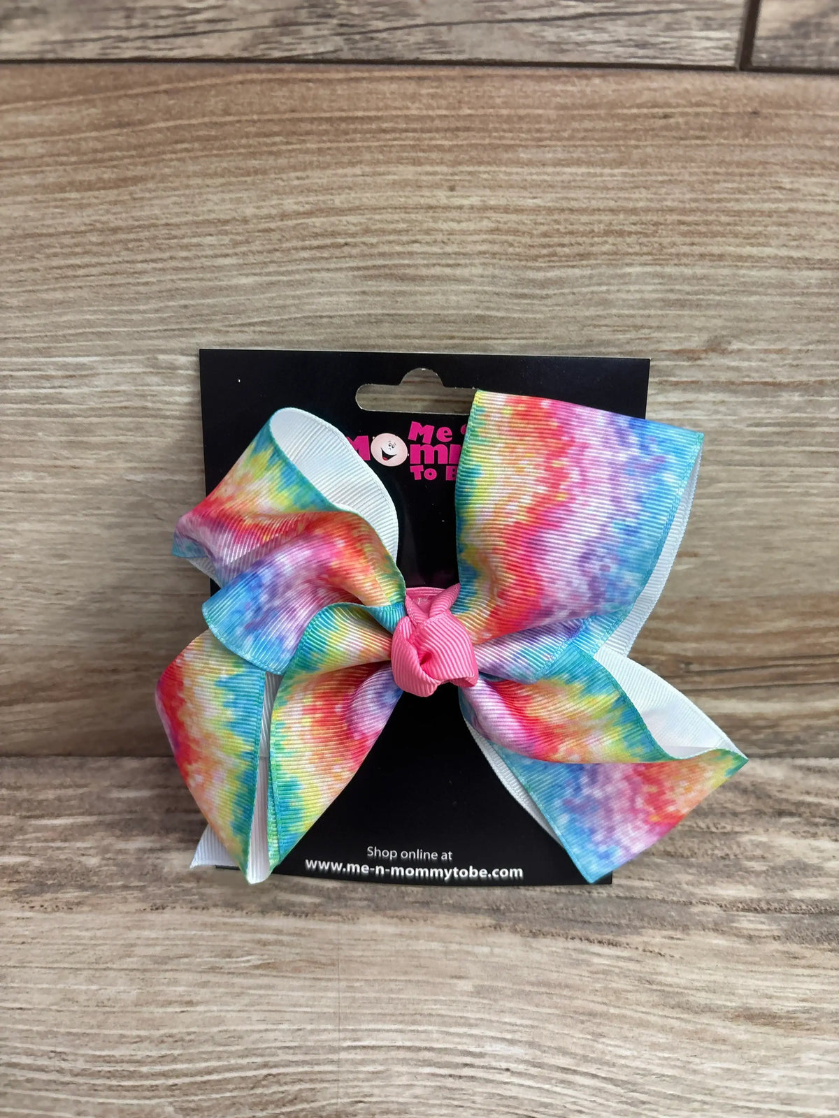 Front view of large rainbow grosgrain hair bow showcasing pink tied center and white layered backing on a black display card over a wood surface