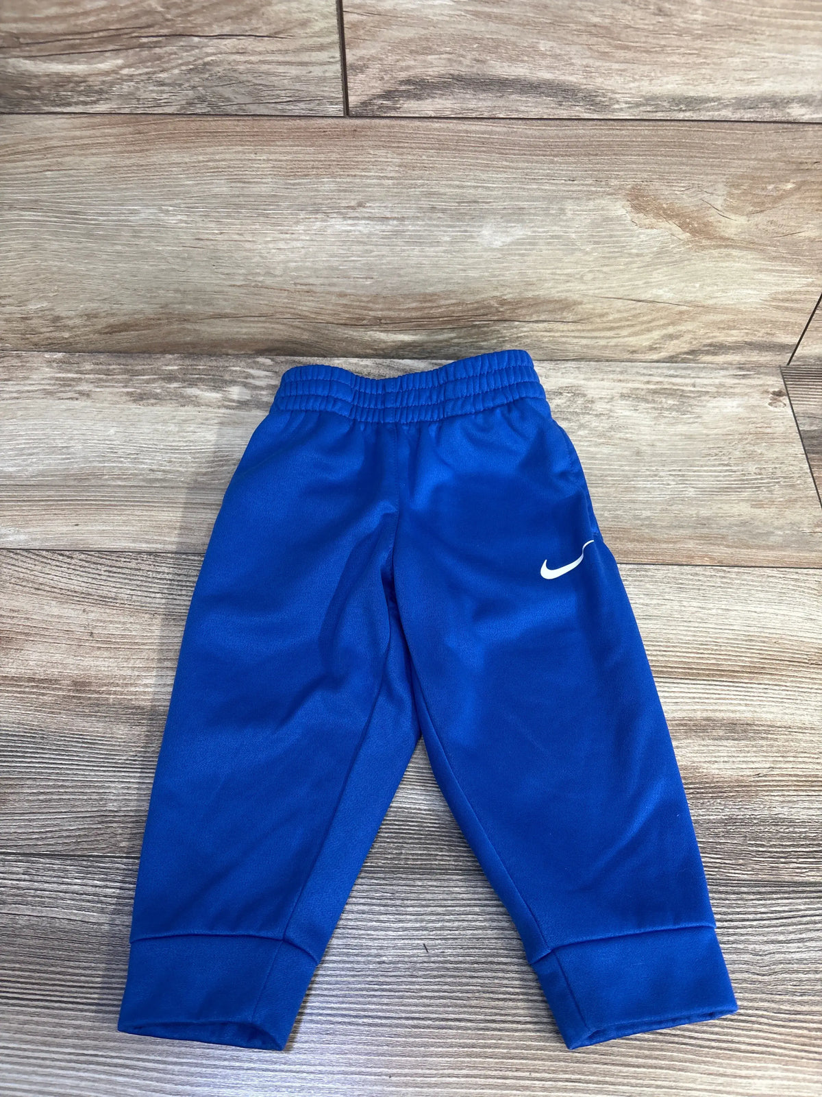Front view of Nike Therma-Fit joggers showcasing blue fabric, elastic waistband, tapered legs, and white swoosh logo on a wood floor