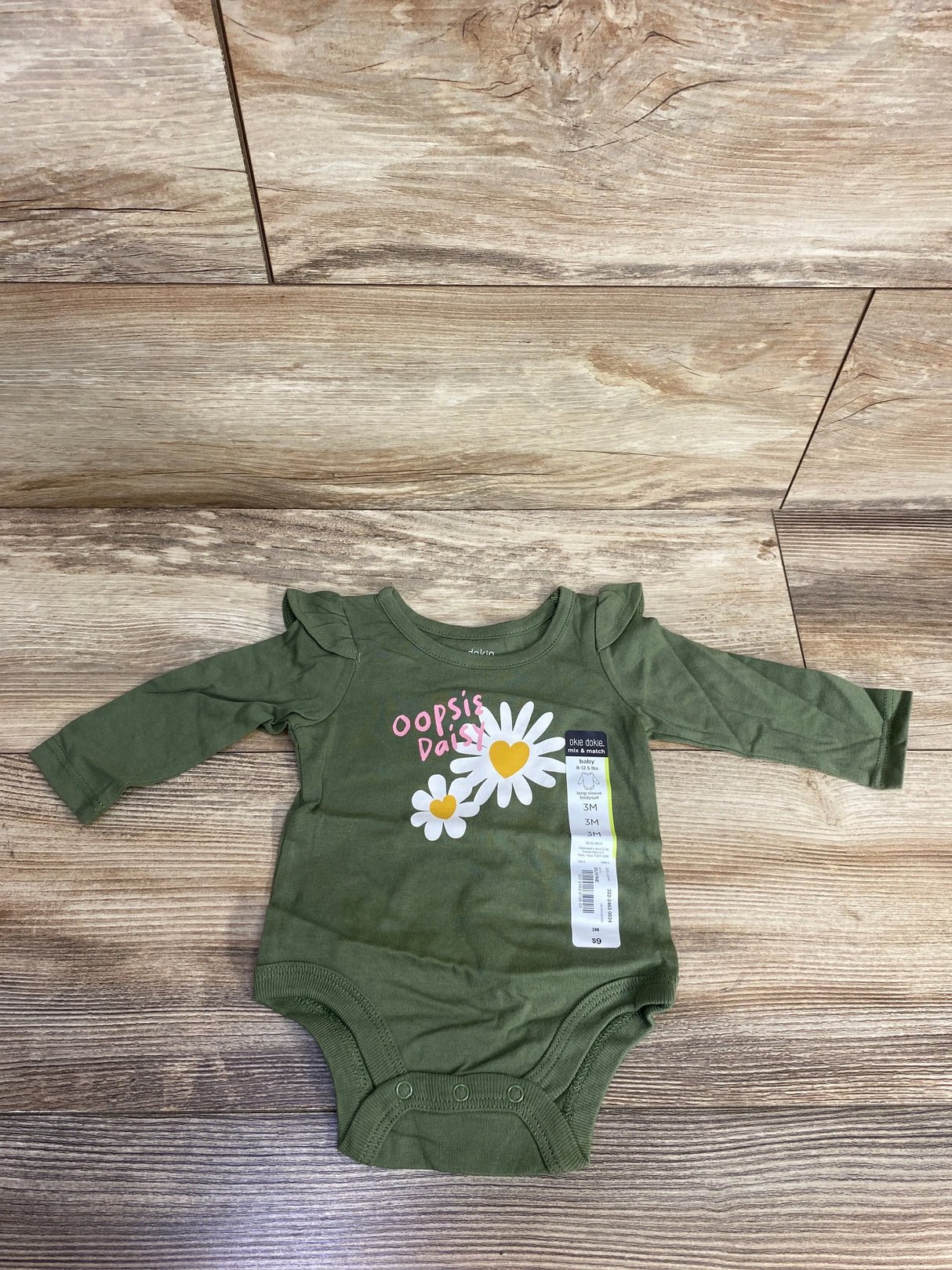 Flat lay of Okie Dokie olive green 3M long-sleeve bodysuit showing ruffled puff shoulders and daisy graphic reading oopsie daisy on wood background