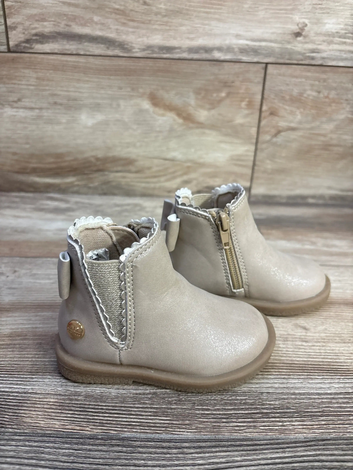 Front-side view of beige Nicole Miller toddler ankle boots showing scalloped trim, side zipper and back bow tab on wood floor
