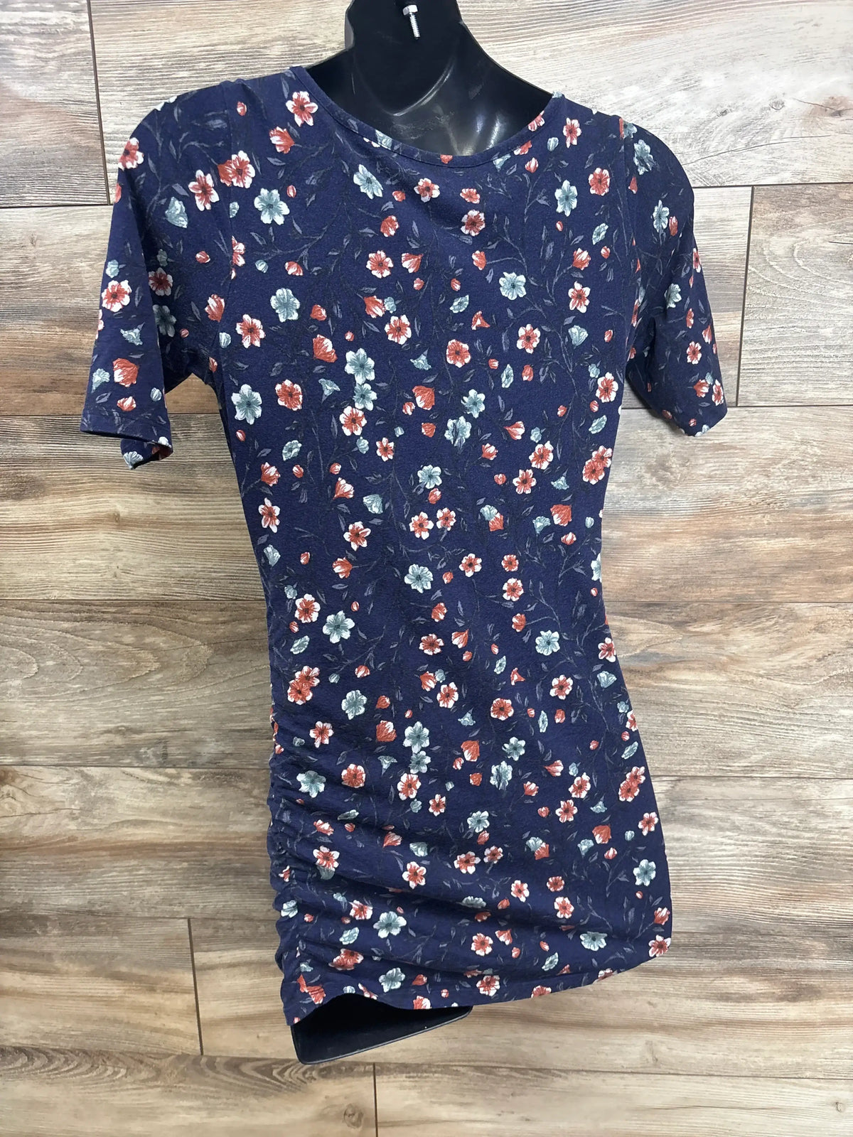 Front view of Motherhood Maternity navy floral ruched shirt showcasing short sleeves and side ruching on a wooden floor
