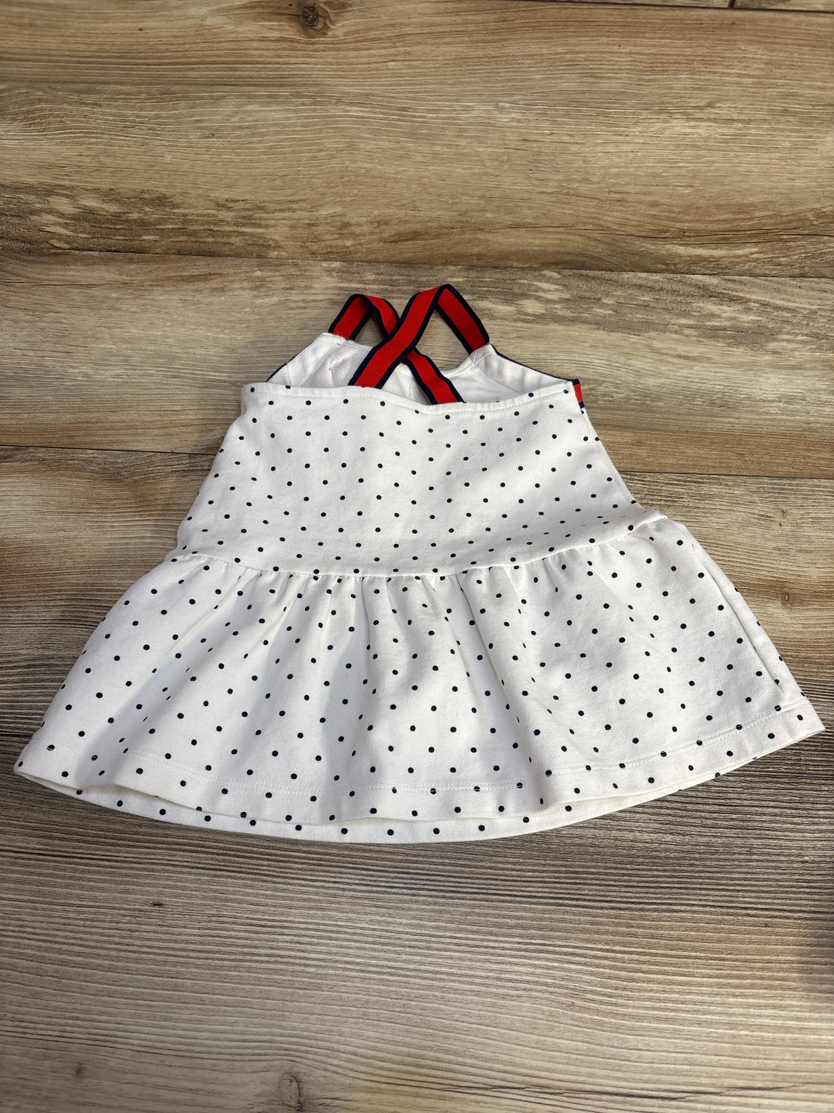 Flat lay of white dot ponte baby dress with black polka dots, gathered skirt, and red and navy crisscross straps on wood