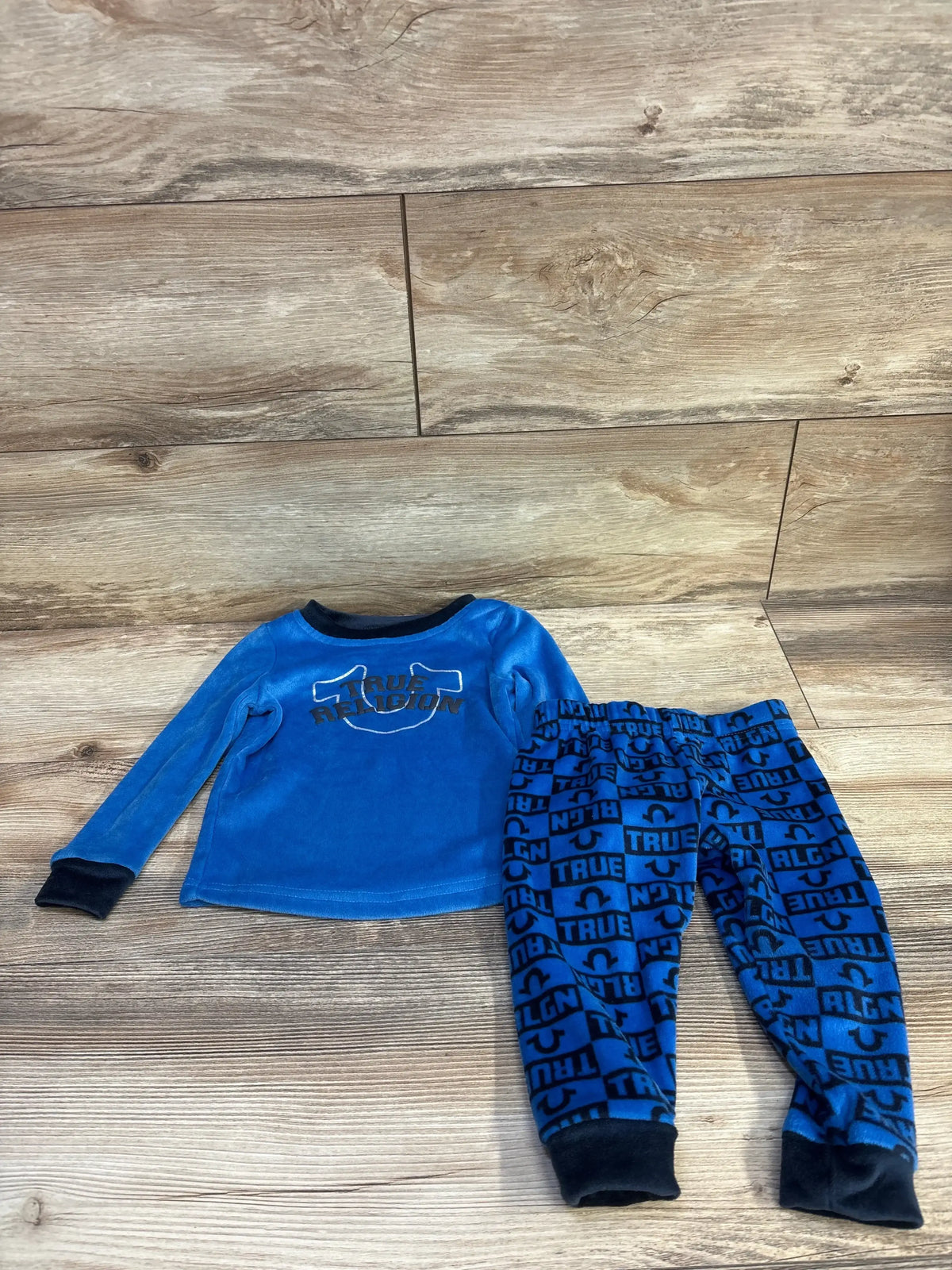 Flat lay of blue True Religion 2-piece fleece pajama set with logo top and matching logo-print pants on wood floor