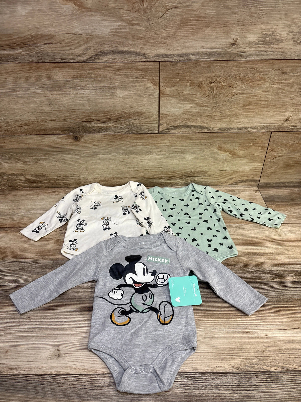 Top view of three Disney Baby Mickey Mouse long sleeve bodysuits in grey, white, and mint patterns, size 6/9 months