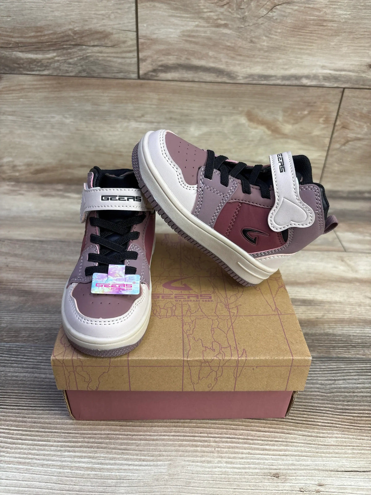 Three-quarter view of mauve and blush Geers high-top toddler sneakers showing Velcro strap, black laces, and textured sole on a cardboard shoebox and wood background