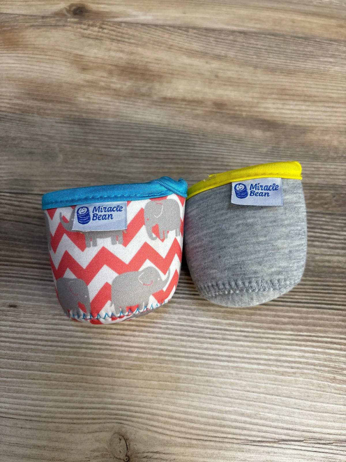 Front view of two Miracle Bean neoprene bottle sleeves, gray with yellow trim and coral chevron with elephant print on wood