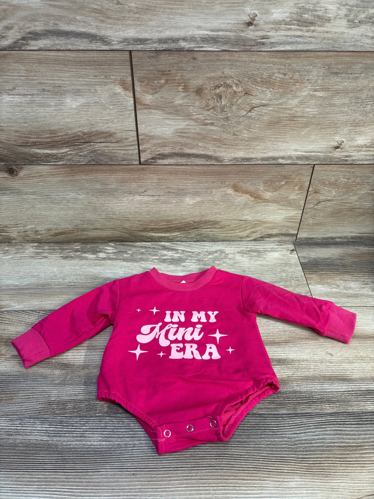 Flat-lay front view of hot pink long-sleeve newborn bodysuit showcasing IN MY MINI ERA print and snap closures on light wood background