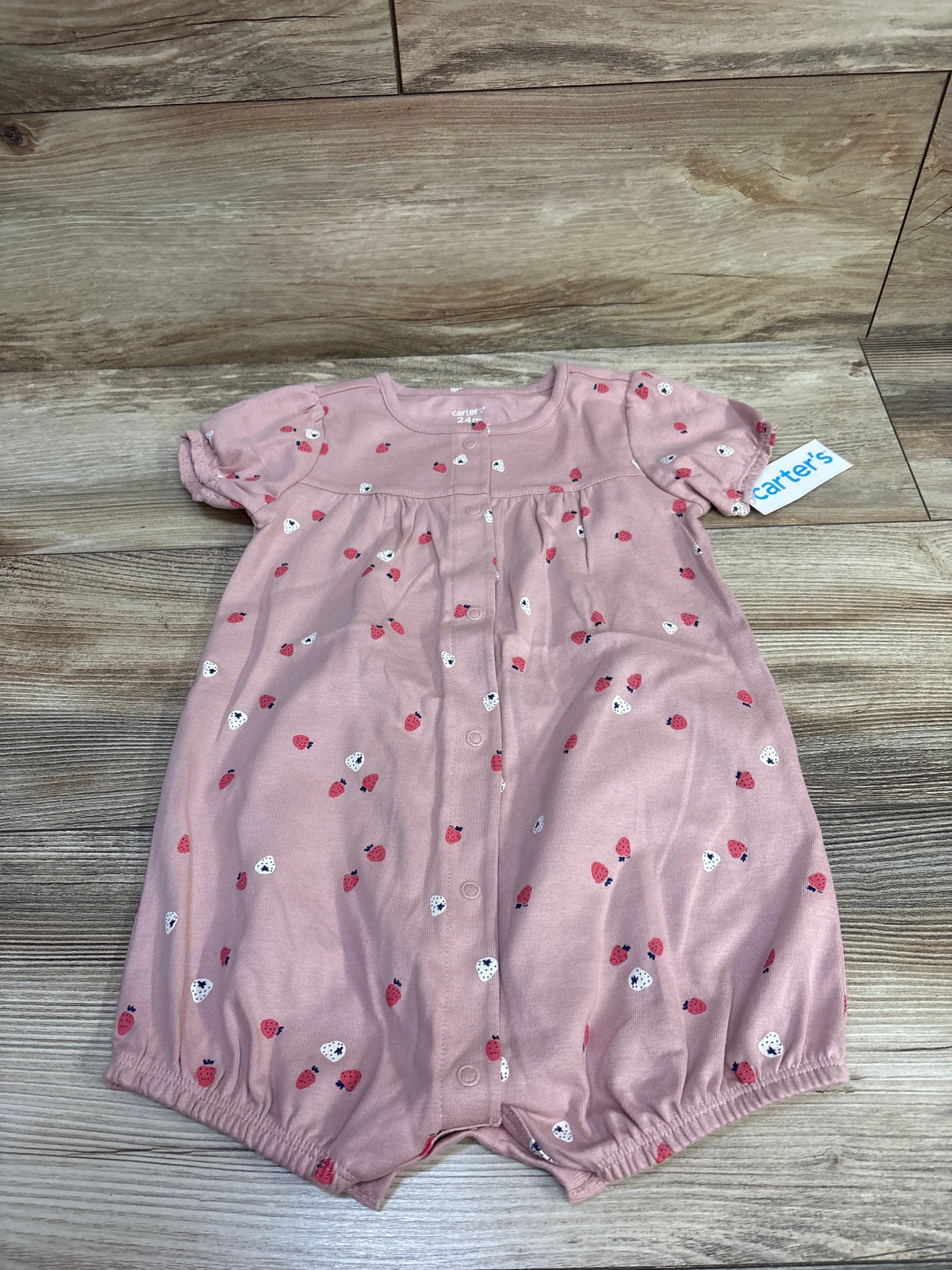 Front view of Carter’s pink 24m strawberry print shortie romper with button front and elastic leg openings on wood