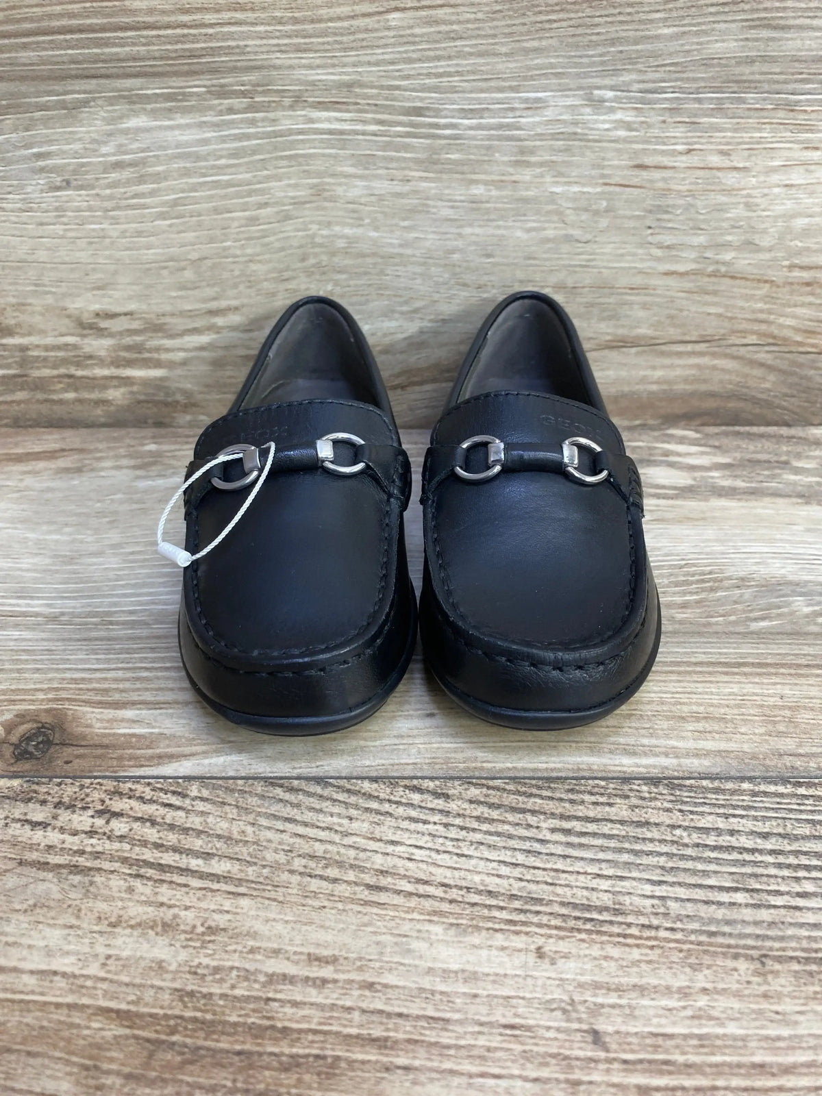 Front view of GEOX toddler black leather loafers showcasing silver chain detail on light wood background
