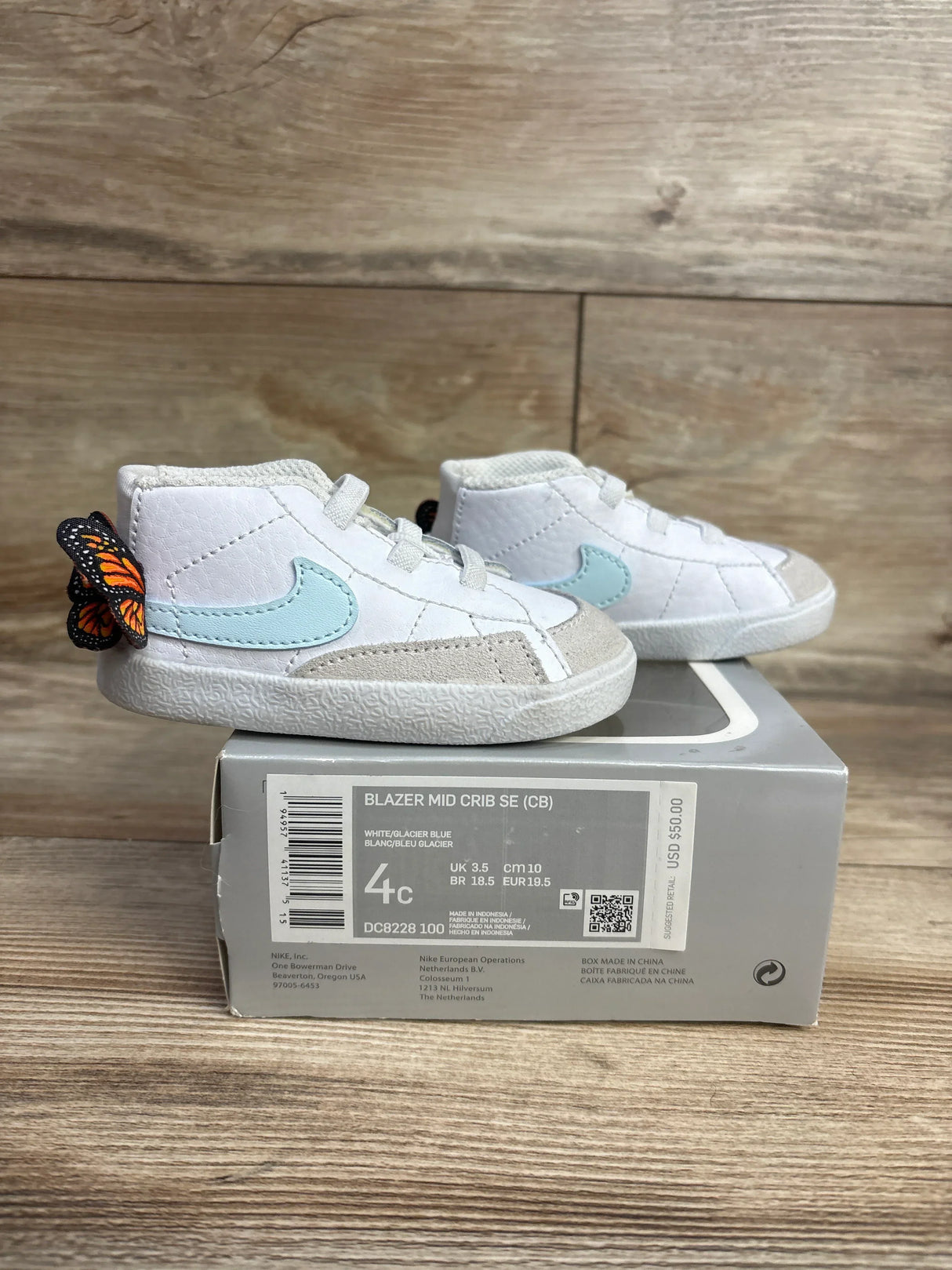 White and glacier blue Nike Blazer Mid Crib SE baby shoes with butterfly detail on gray box on wooden floor