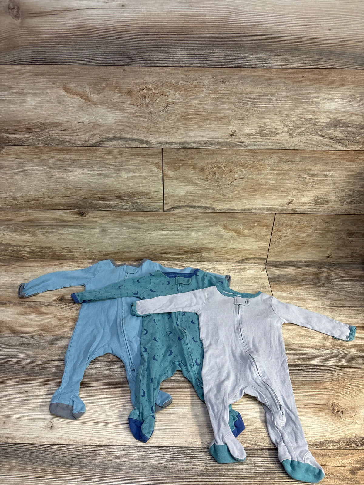 Flat lay of three Cloud Island blue footed sleepers (solid, moon-print, light heather) showing front zippers on wood background