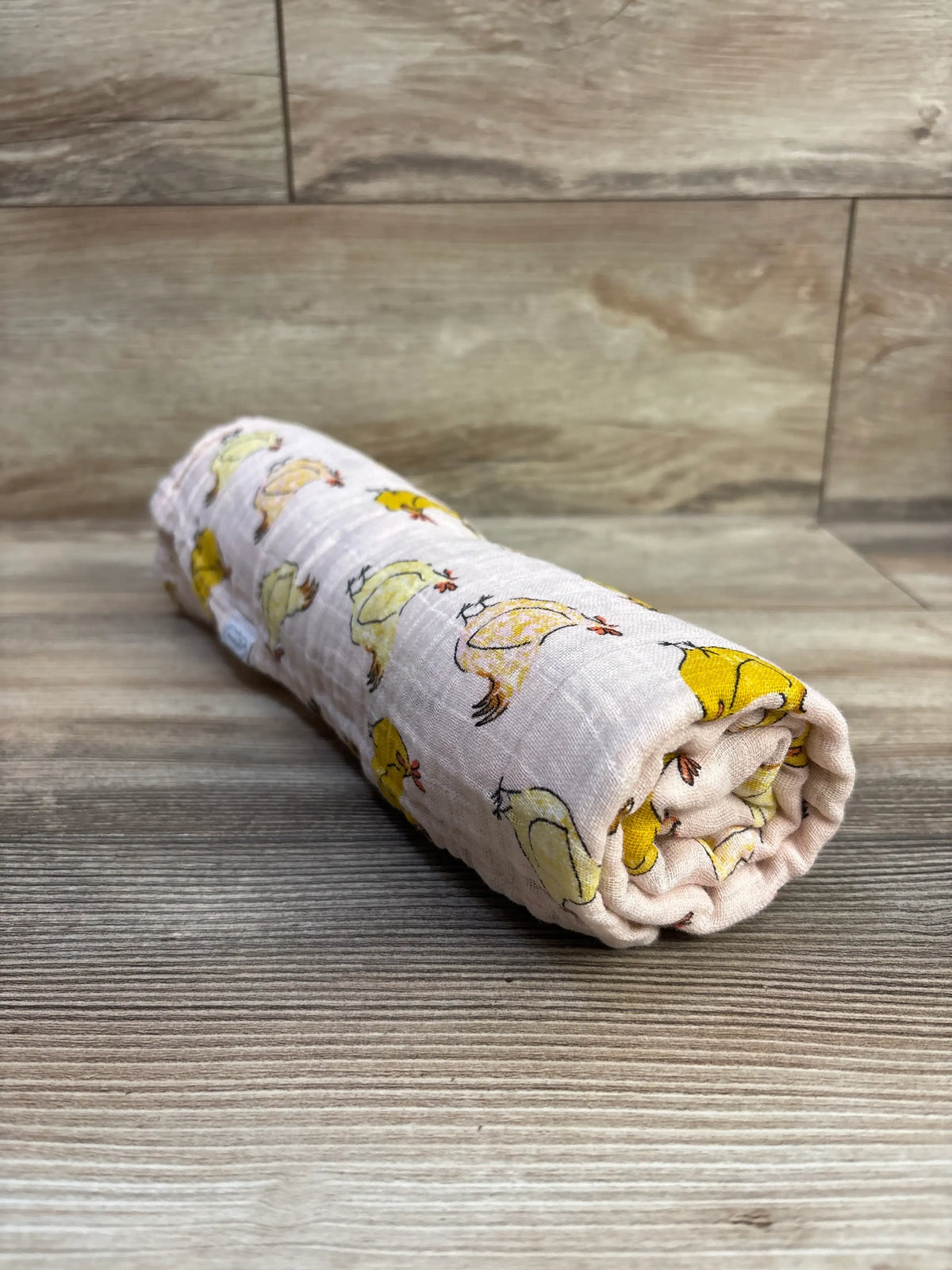 Rolled view of a pink chicken muslin swaddle blanket showcasing its chicken print with a wood-look floor and wall background