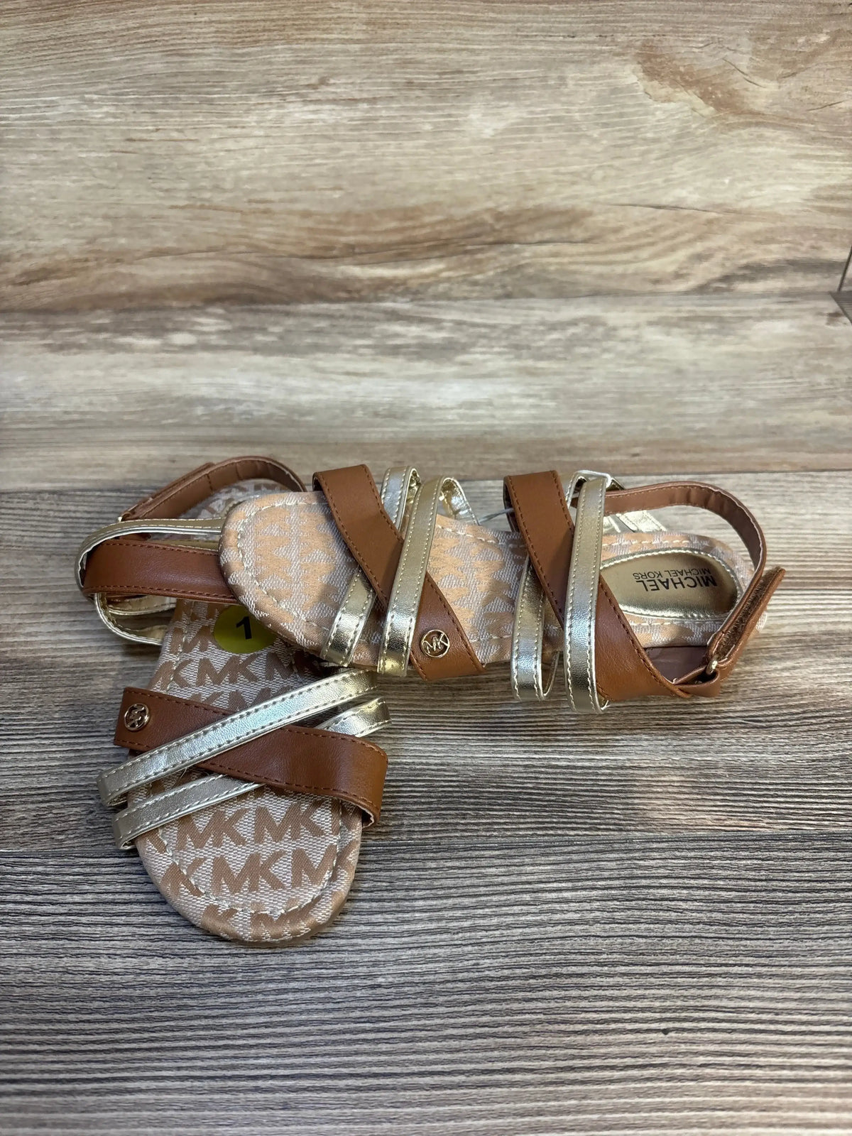 Brown and gold toddler sandals with MK logo, strappy design on wood background