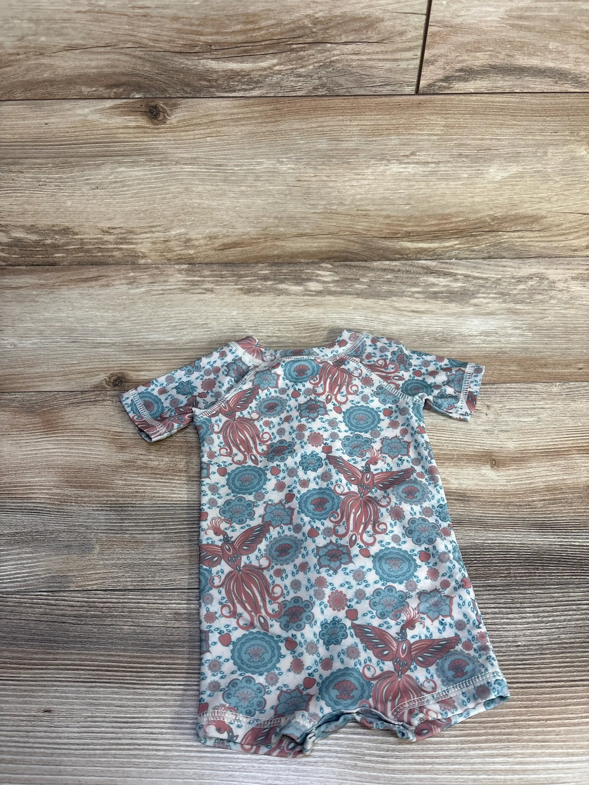 Flat lay of 0-3m rash guard shortie swimsuit in ivory with teal and coral octopus print on light wood background