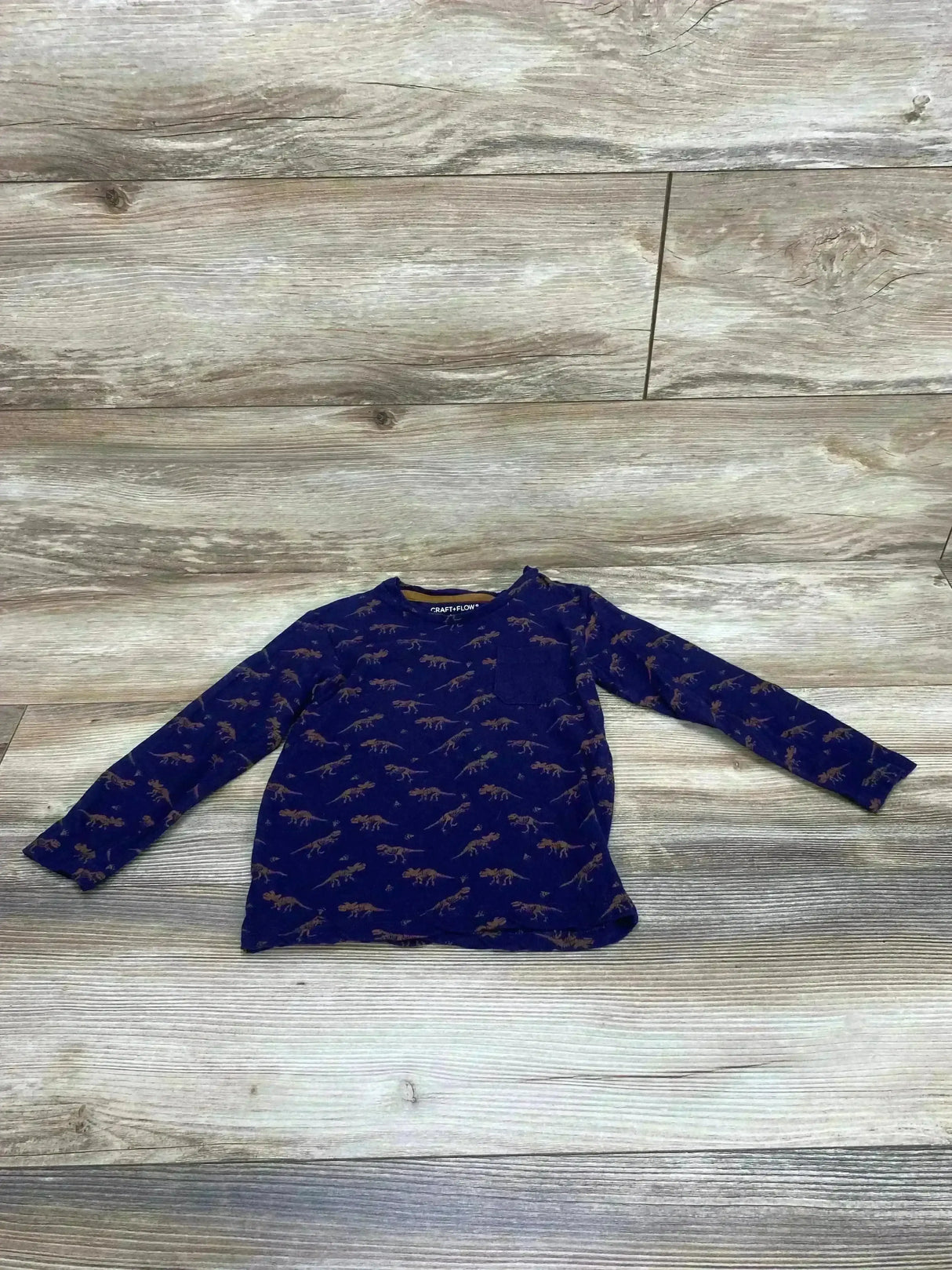Flat lay of navy long-sleeve toddler shirt (size 4T) showcasing all-over brown dinosaur print and chest pocket on light wood floor