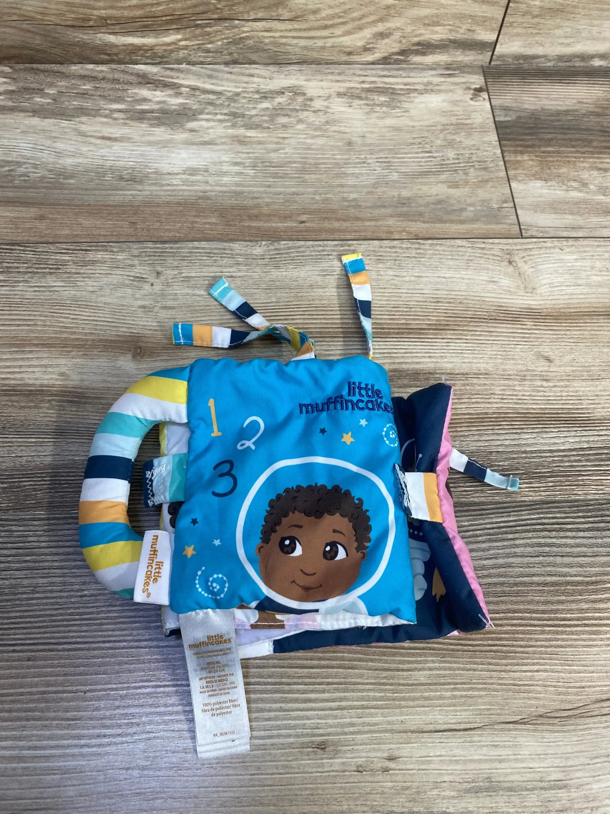 Top-down view of Little Muffincakes Ashton Blue soft baby book with astronaut-face cover, striped handle and fabric tags on floor