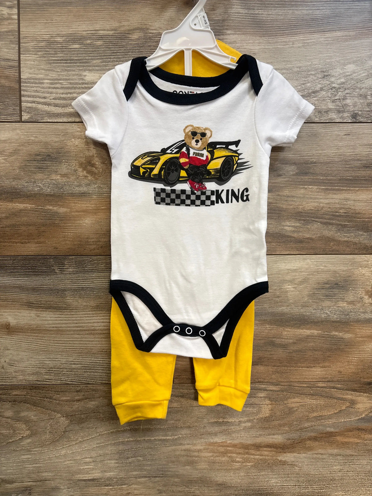 Front view of white short-sleeve baby bodysuit with black trim and racecar bear graphic paired with yellow pants on wood background