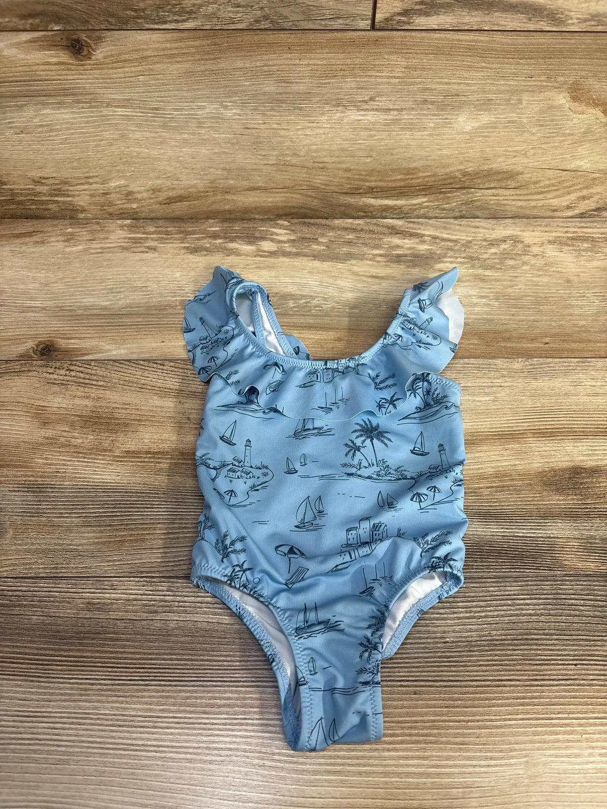 Flat-lay front view of baby blue one-piece swimsuit showcasing beach print and ruffled shoulder straps on wood background