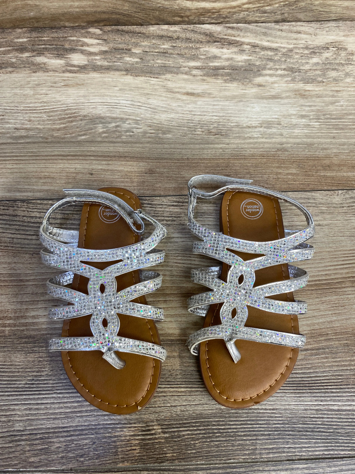 Sparkly silver gladiator sandals with brown soles on a wooden floor