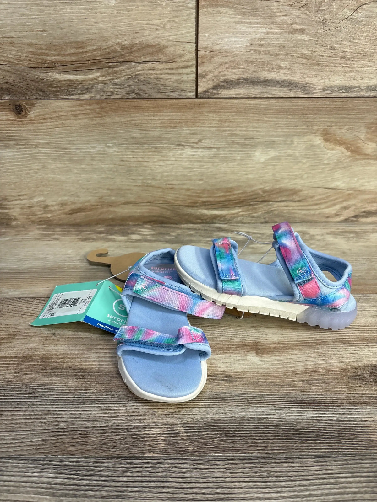 Front view of toddler blue Lumos light-up sandals with tie-dye Velcro straps and clear light-up soles on wood background