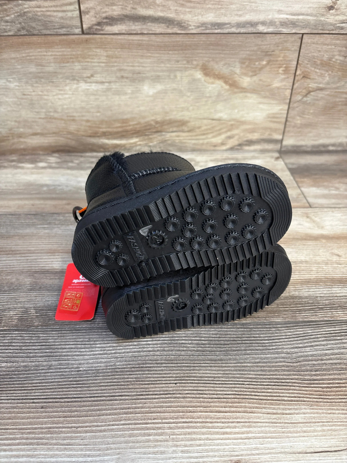 Black toddler winter boots with textured rubber soles on wood floor, red tag attached