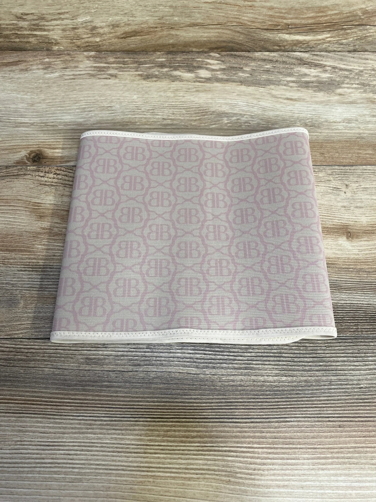 Top-down of Belly Bandit postpartum belly wrap, beige with pink BB monogram, folded to show elastic trim on light wood background