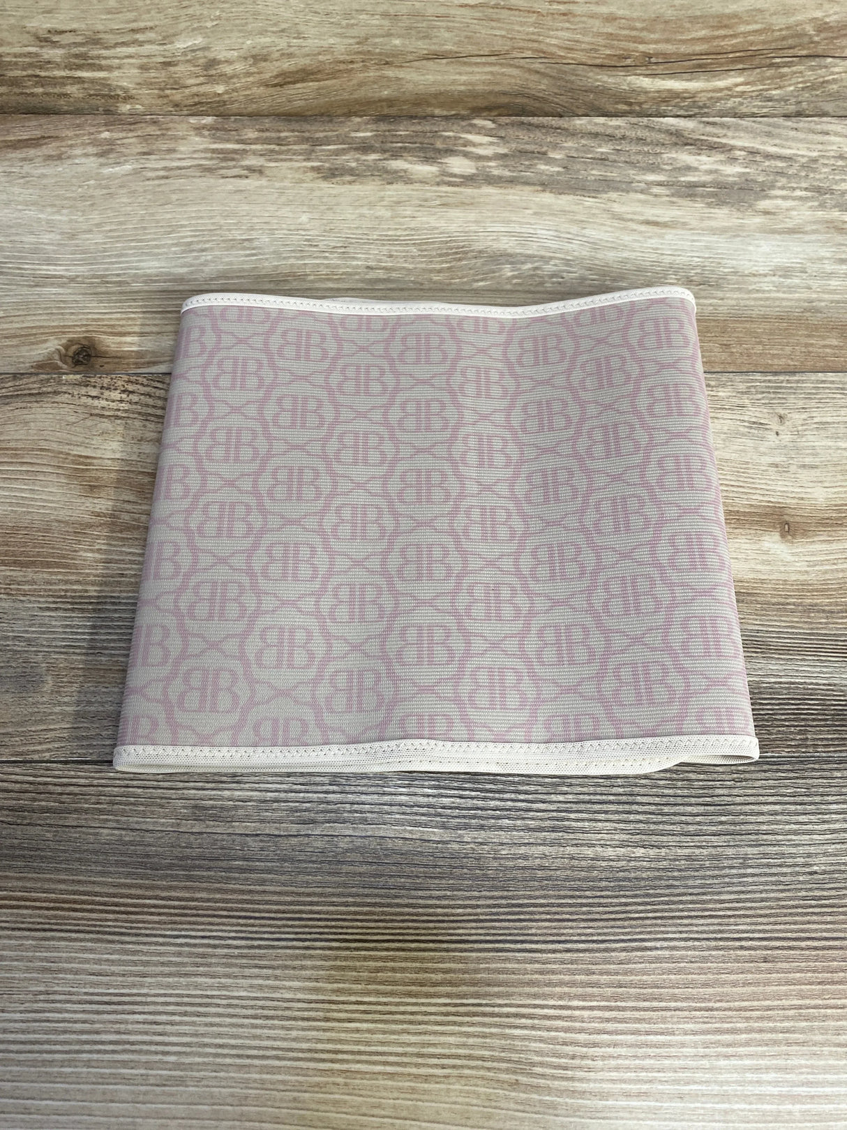 Top-down of Belly Bandit postpartum belly wrap, beige with pink BB monogram, folded to show elastic trim on light wood background