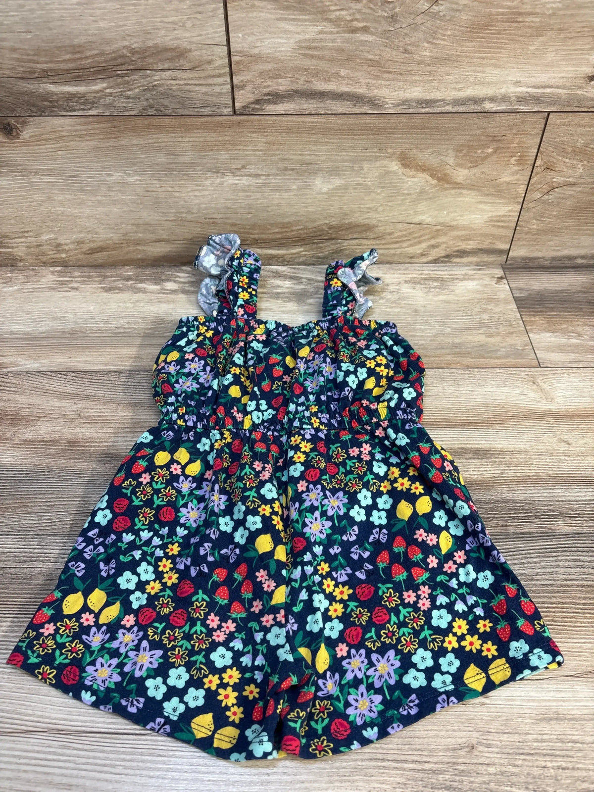 Flat lay of navy sleeveless toddler romper with colorful fruit and floral print and elasticized waist on wood floor background