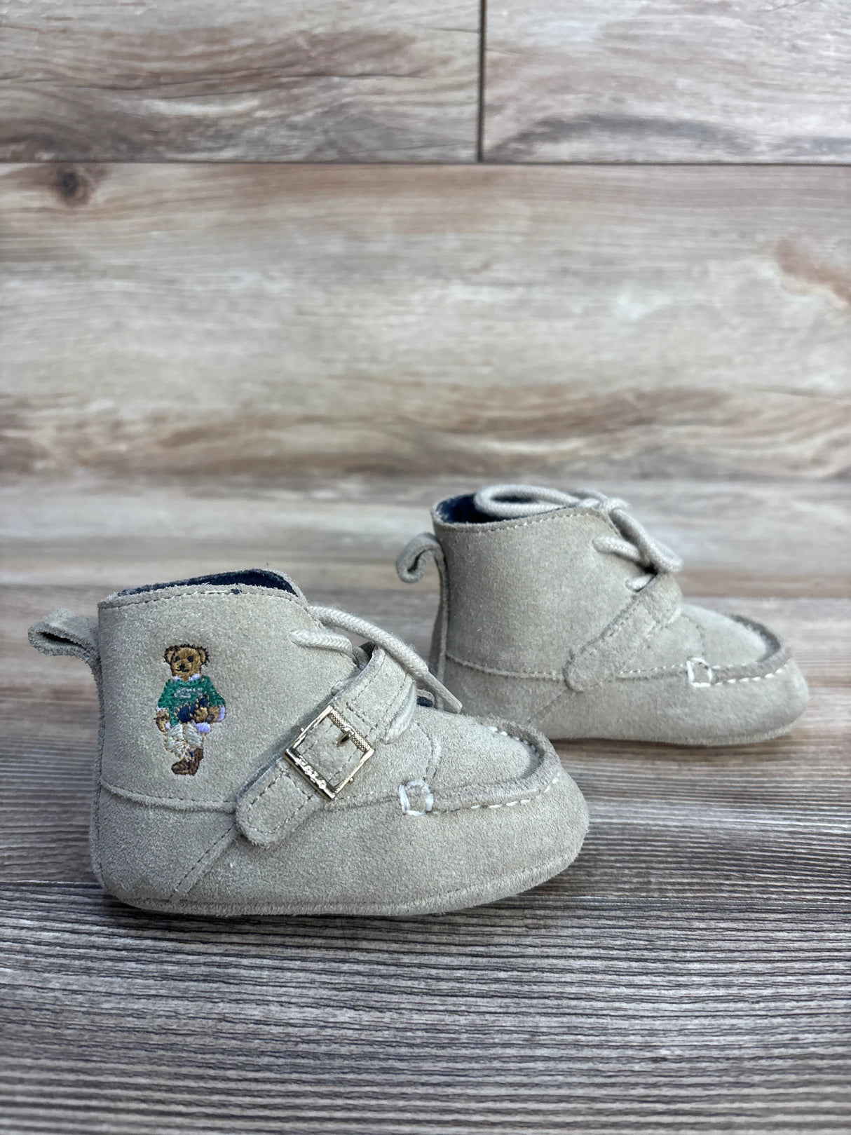 Beige suede baby shoes with teddy bear embroidery and buckle detail on wooden surface