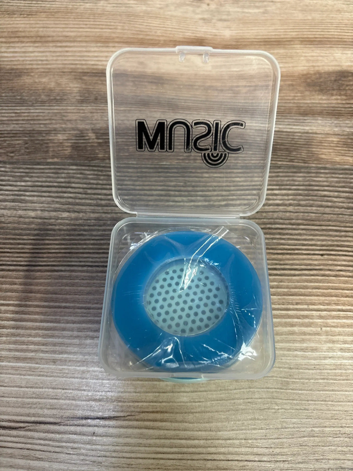 Blue round portable speaker in clear plastic case with 'MUSIC' label on wooden surface