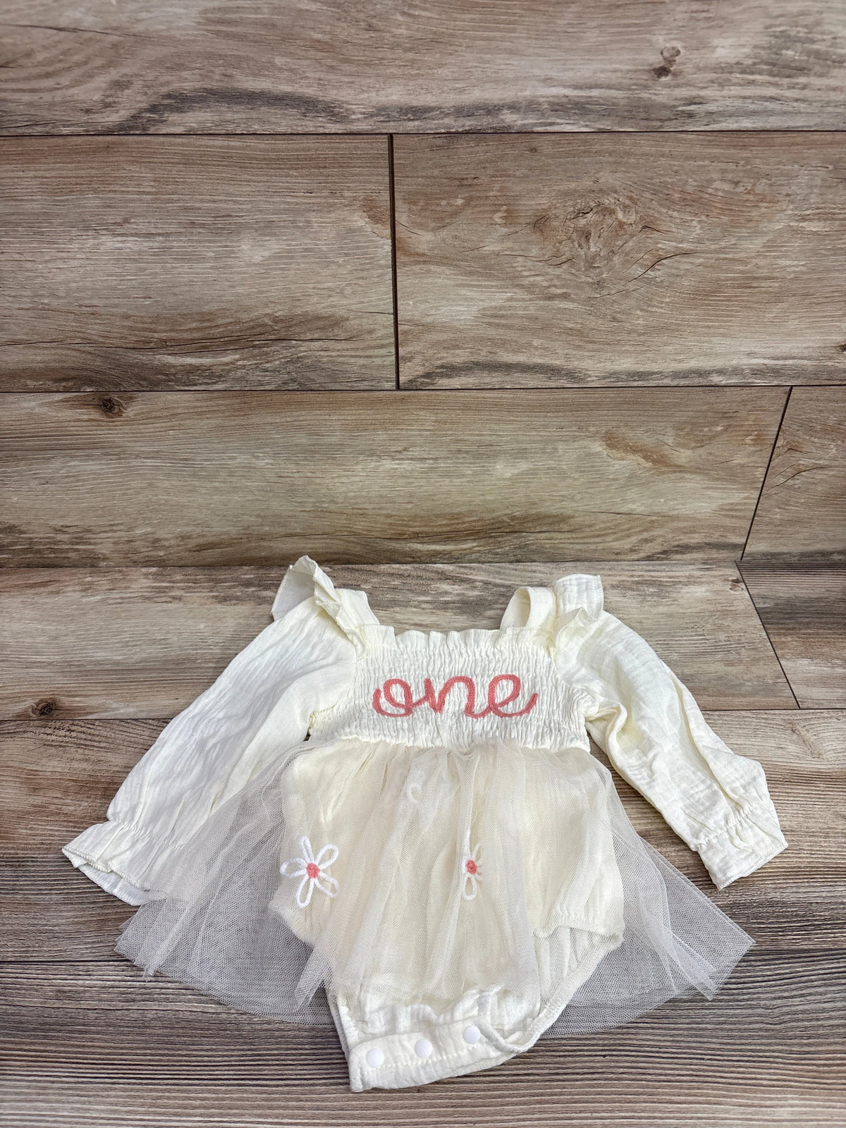 Front view of cream long sleeve smocked first birthday tulle romper with one embroidery and floral accents on wood flooring